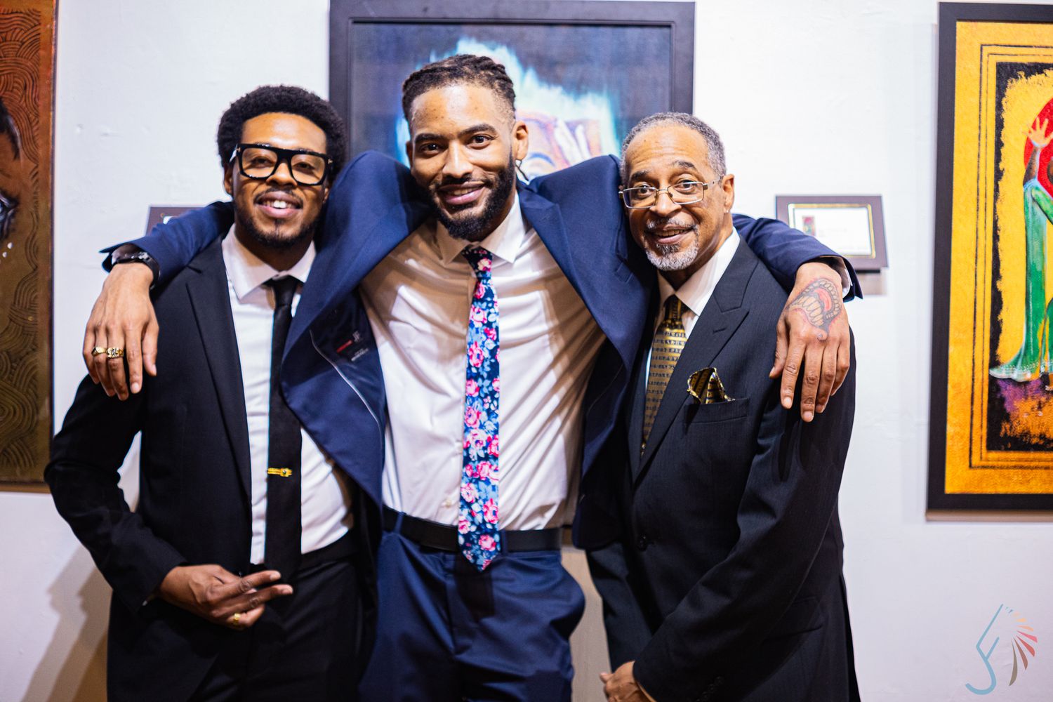 Three well-dressed men in suits smiling and posing together at an art gallery event.