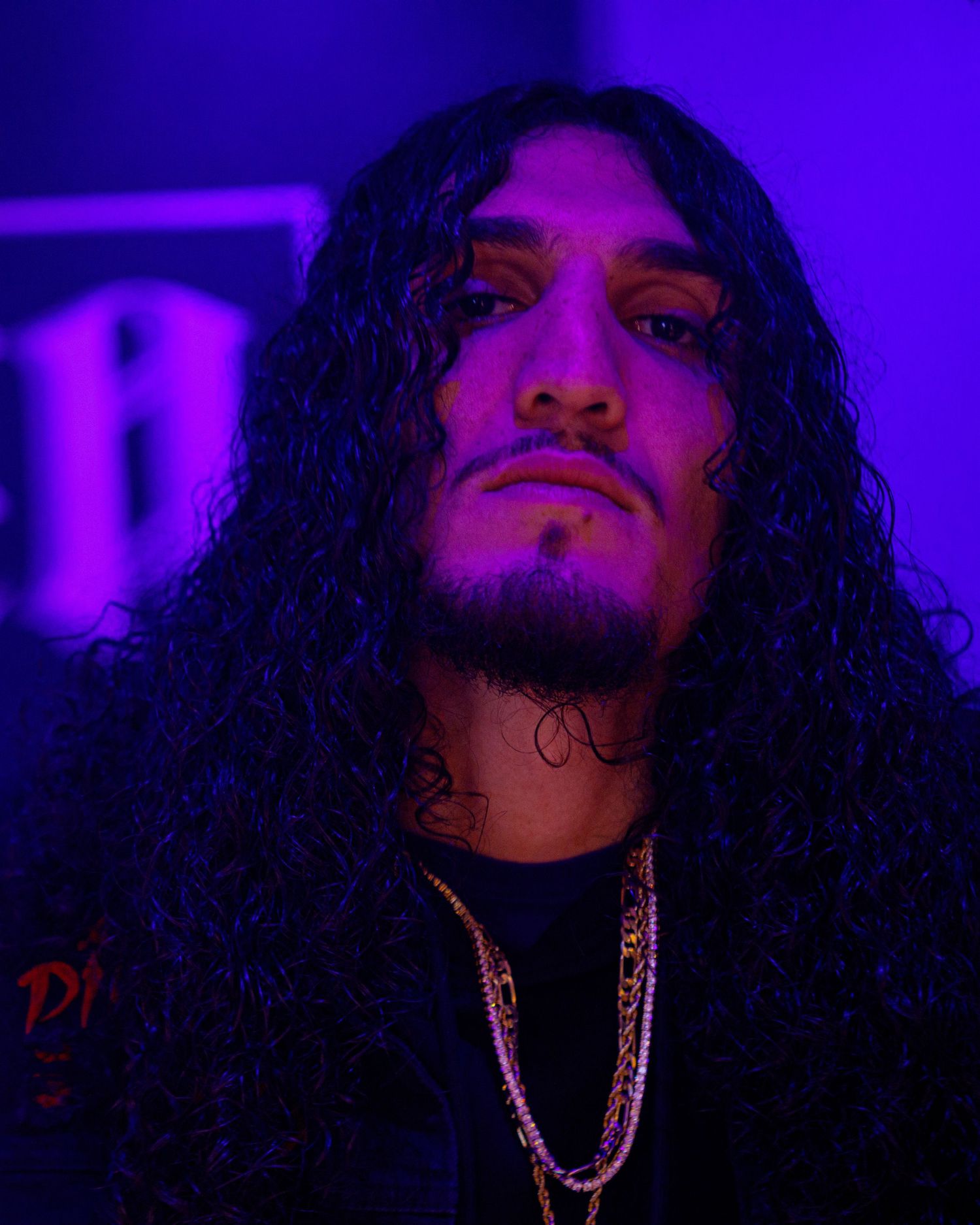 Man with long curly black hair and gold chains under purple lighting.