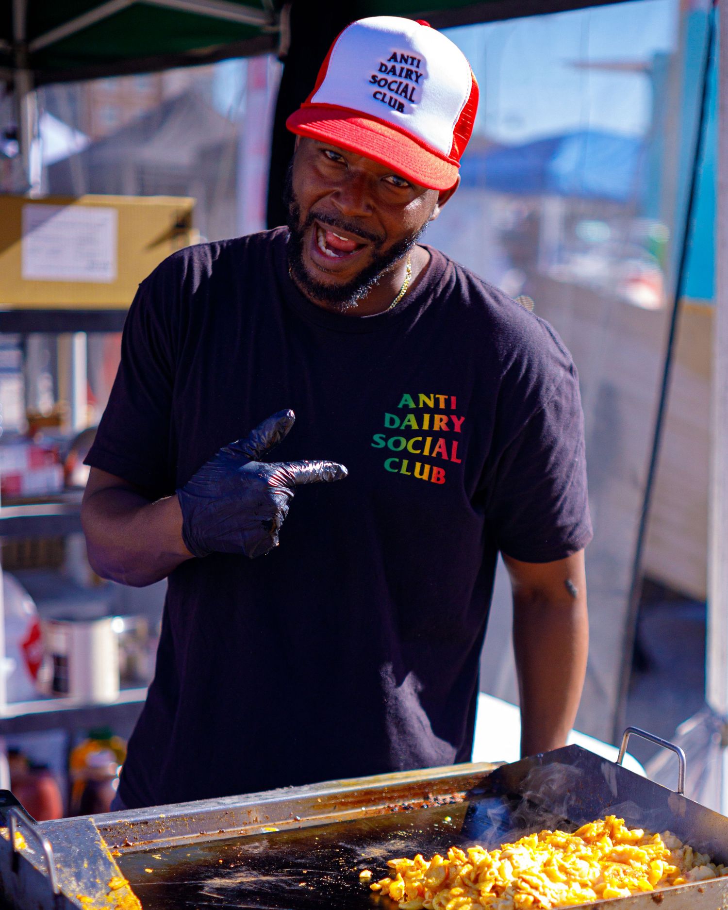 A man wearing an Anti Dairy Social Club shirt and hat cooks mac and cheese on a flat top grill.