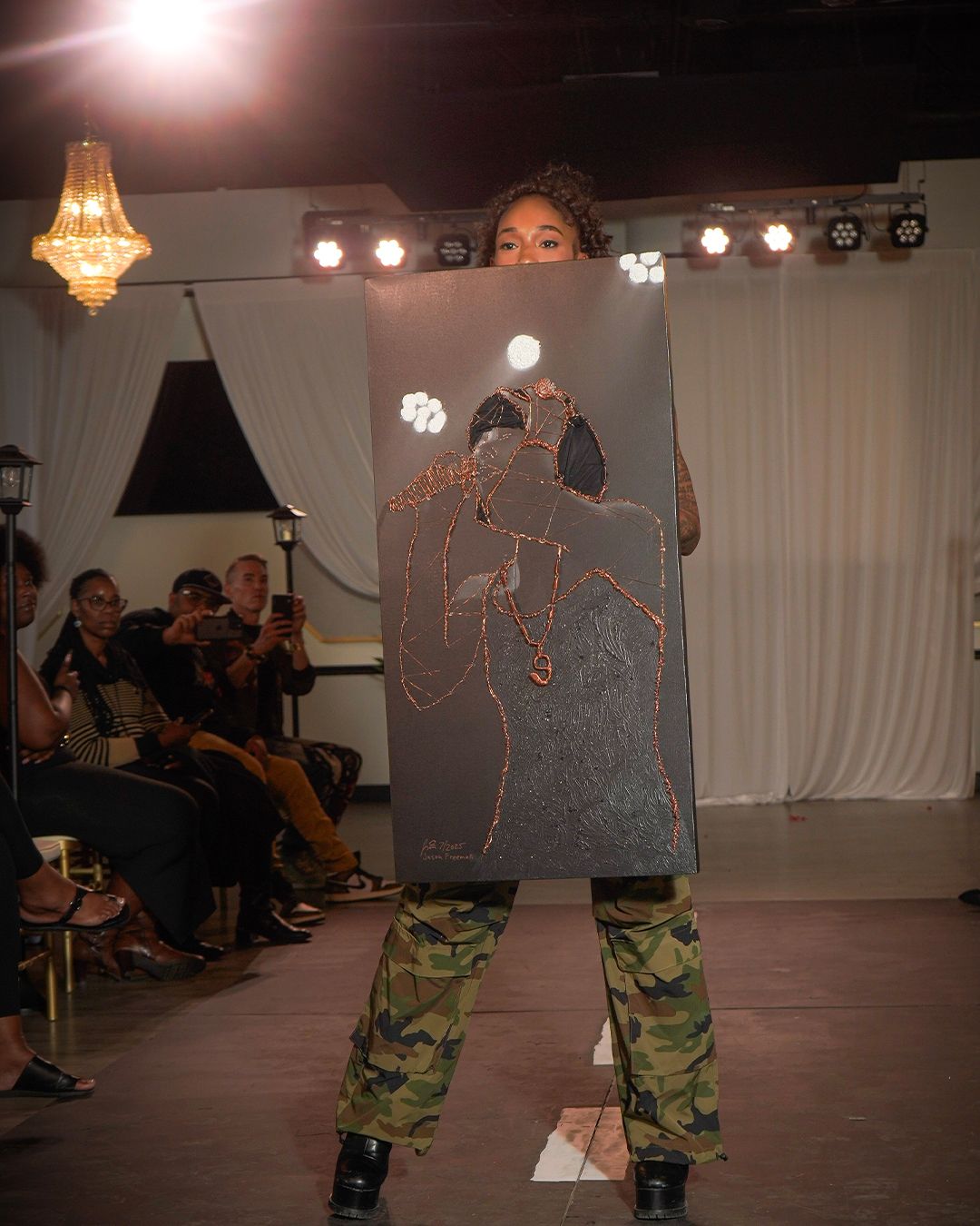 A model in camo pants holds a large dark painting at a fashion show runway event with audience in background.