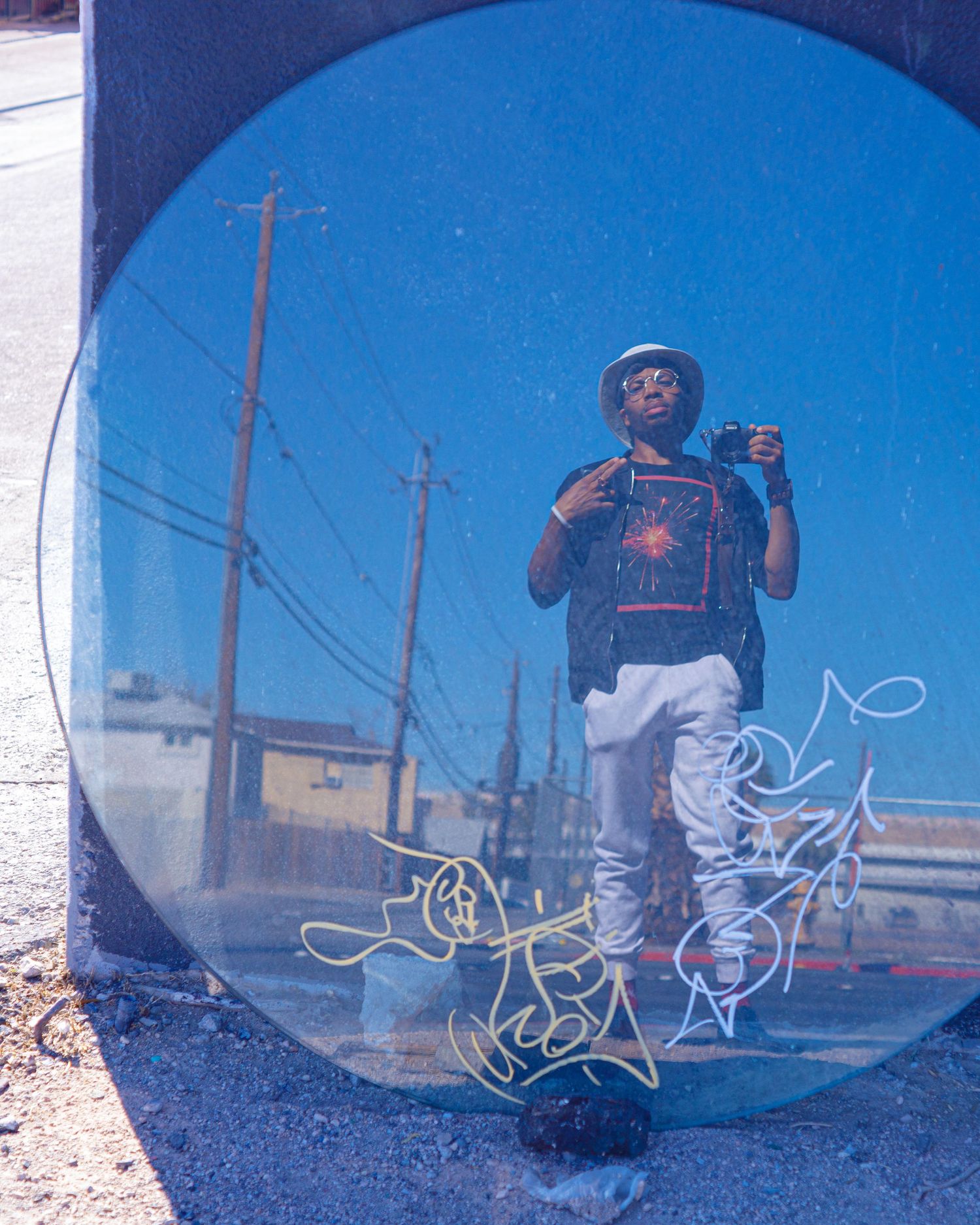 A man poses for a creative selfie reflected in a large circular mirror outdoors with graffiti art.