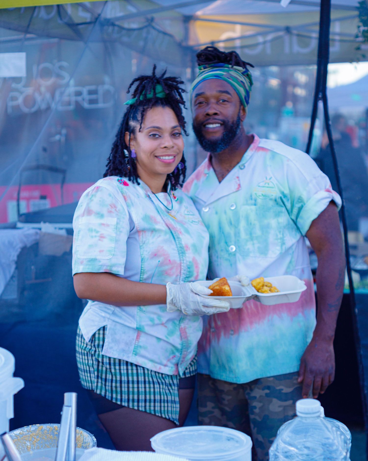 Two vendors in matching tie-dye uniforms holding food at an outdoor festival booth.