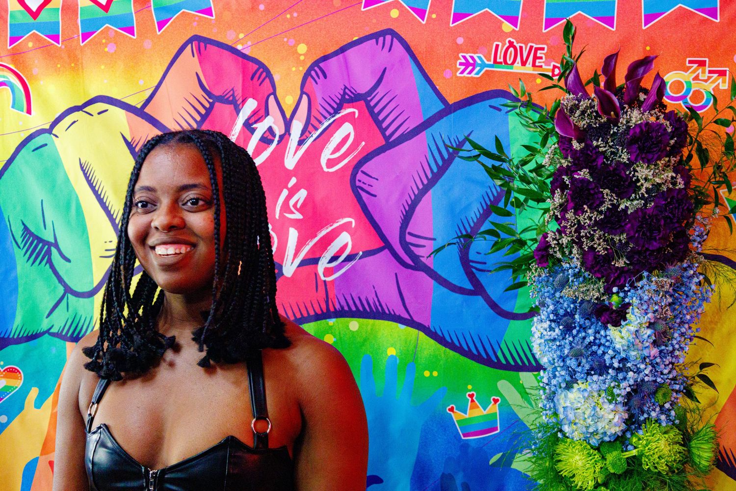 Young woman smiling in front of colorful love is love Pride mural with fist bumps and floral arrangement.