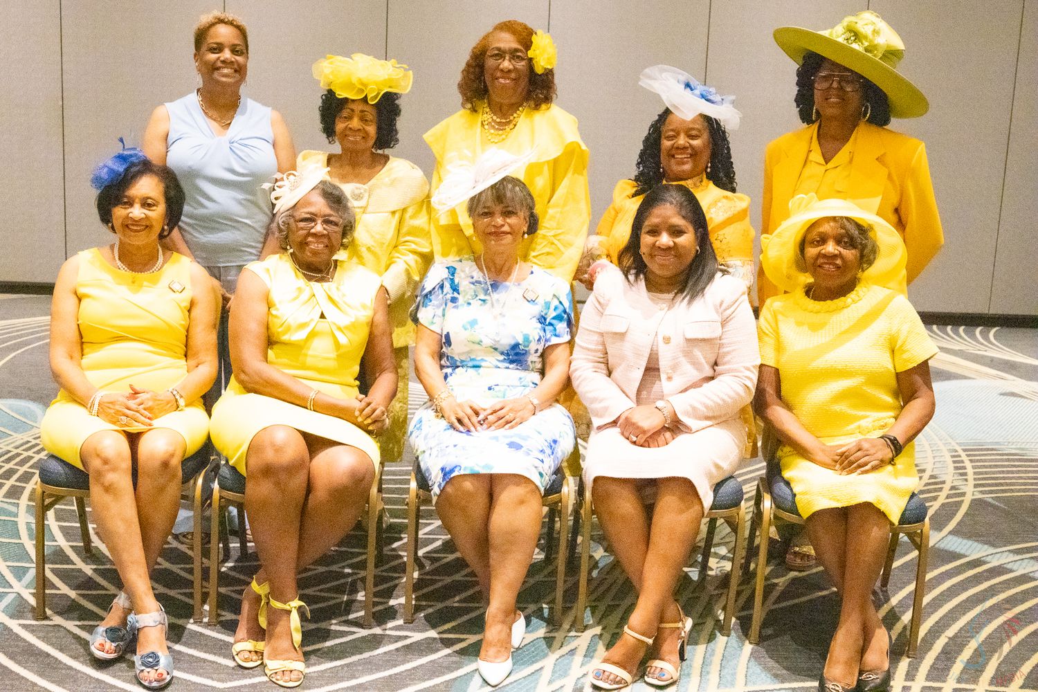 Group of women posing in coordinated yellow outfits and hats for a formal occasion.