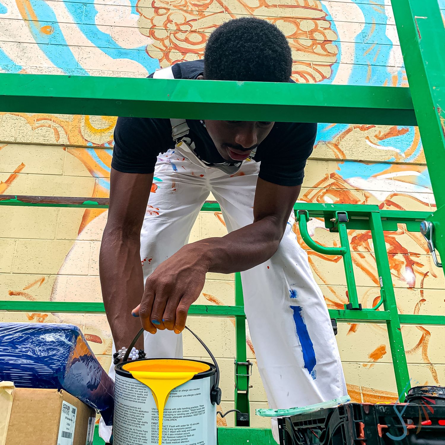 A young Black artist leans over scaffolding, pouring yellow paint while working on a colorful outdoor mural.