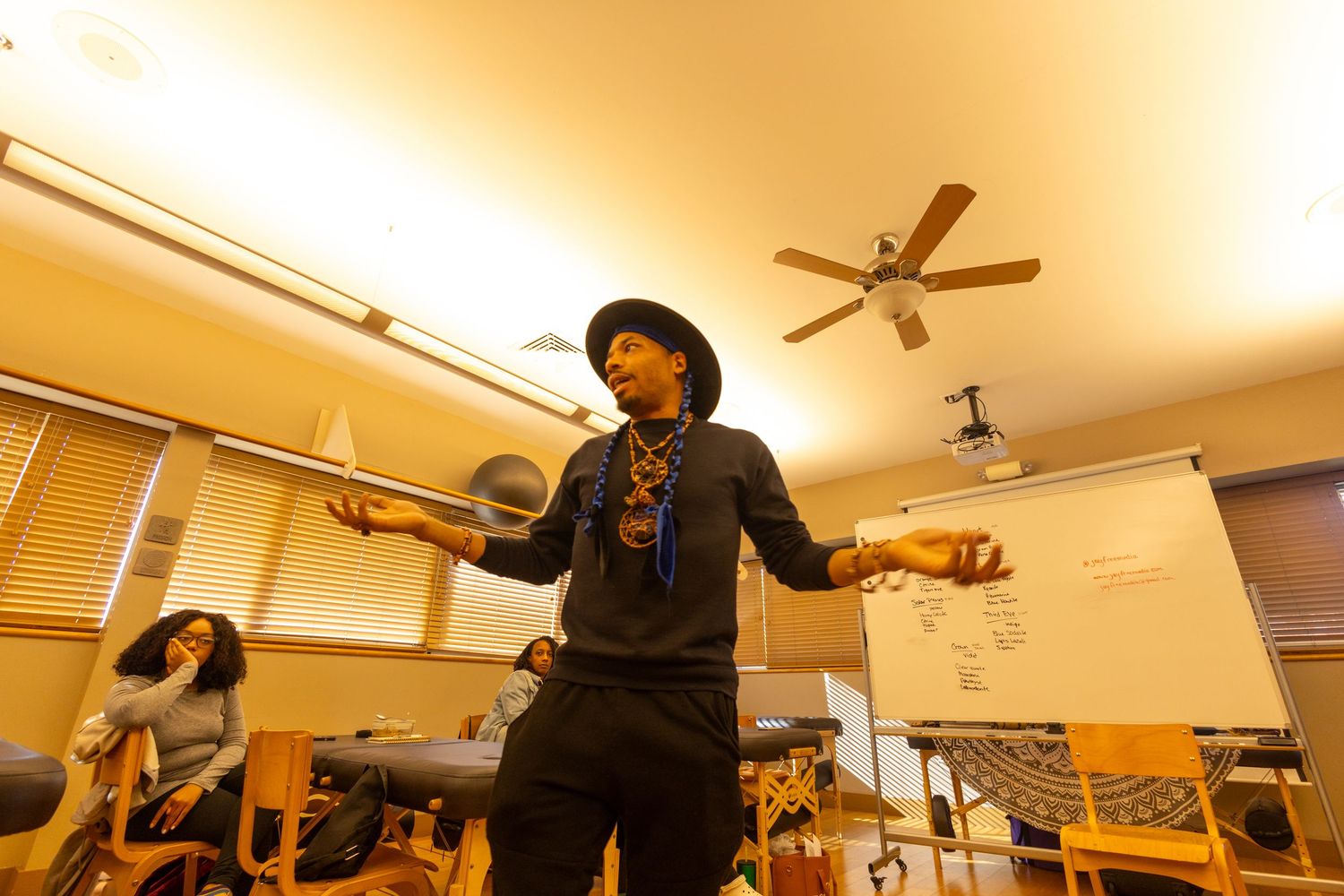 A performer in a black hat gestures expressively in a warmly lit classroom with students and a ceiling fan.