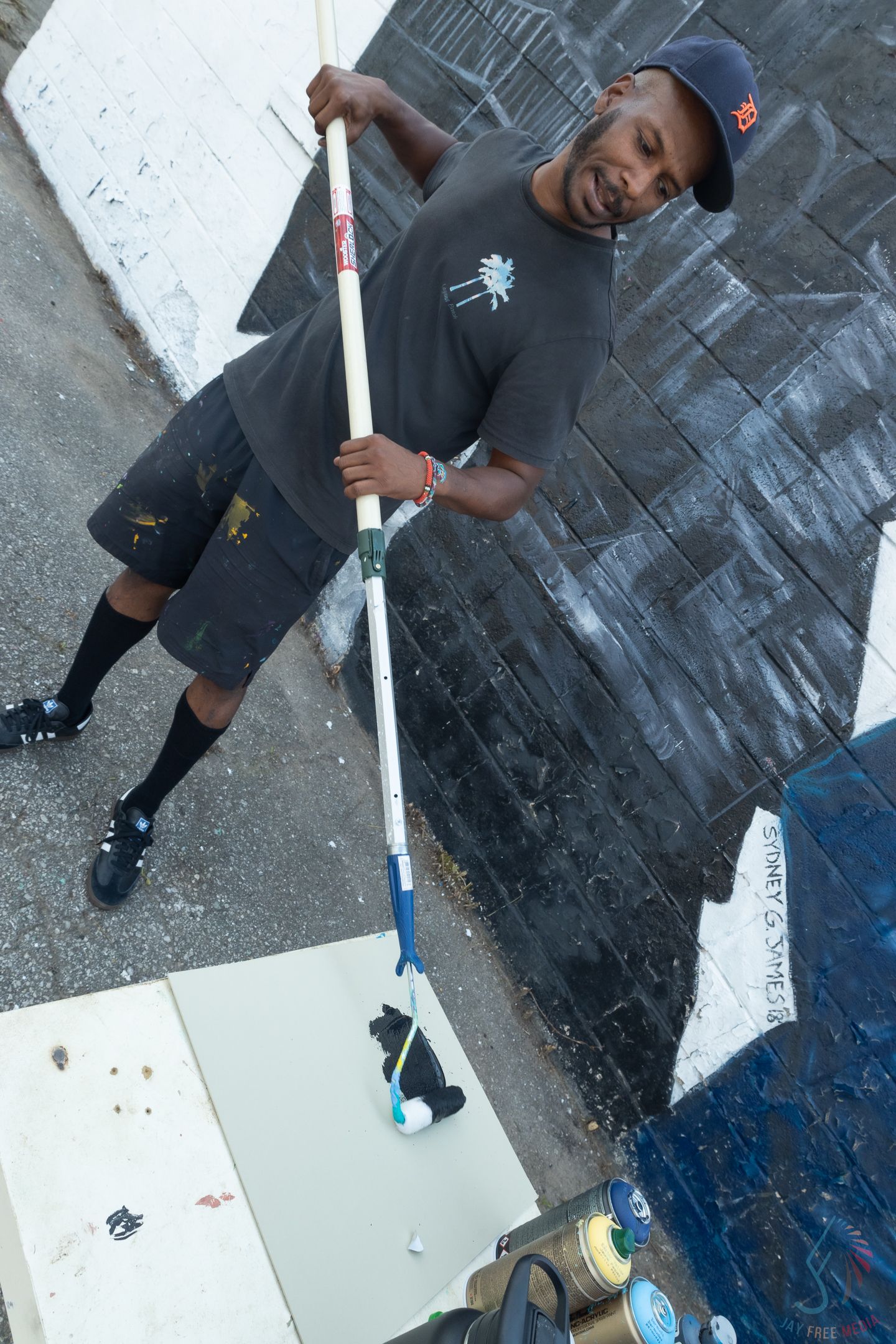 A man in a dark outfit holds a long paint roller near a freshly painted black and white mural on an outdoor wall.