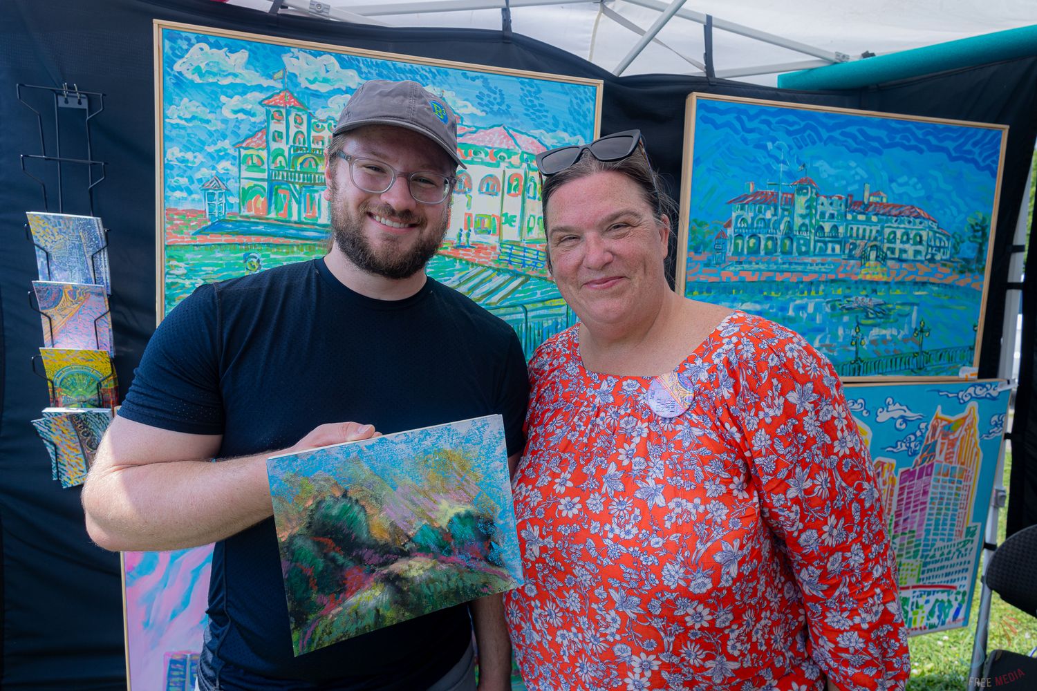 Man and woman pose together holding a colorful landscape painting at an outdoor art festival booth.