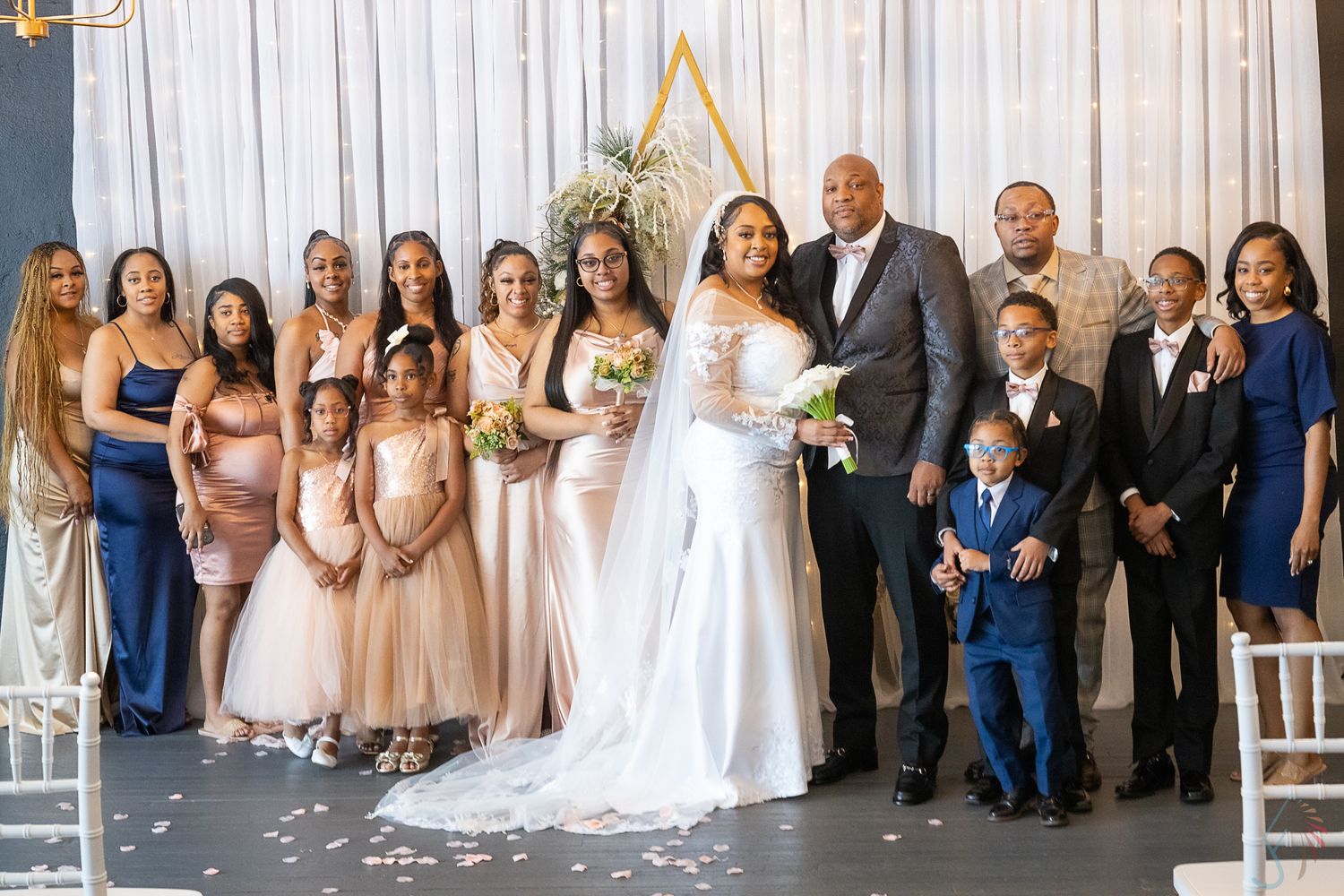 Wedding party group photo with bride, groom, bridesmaids, groomsmen, and children at elegant ceremony.
