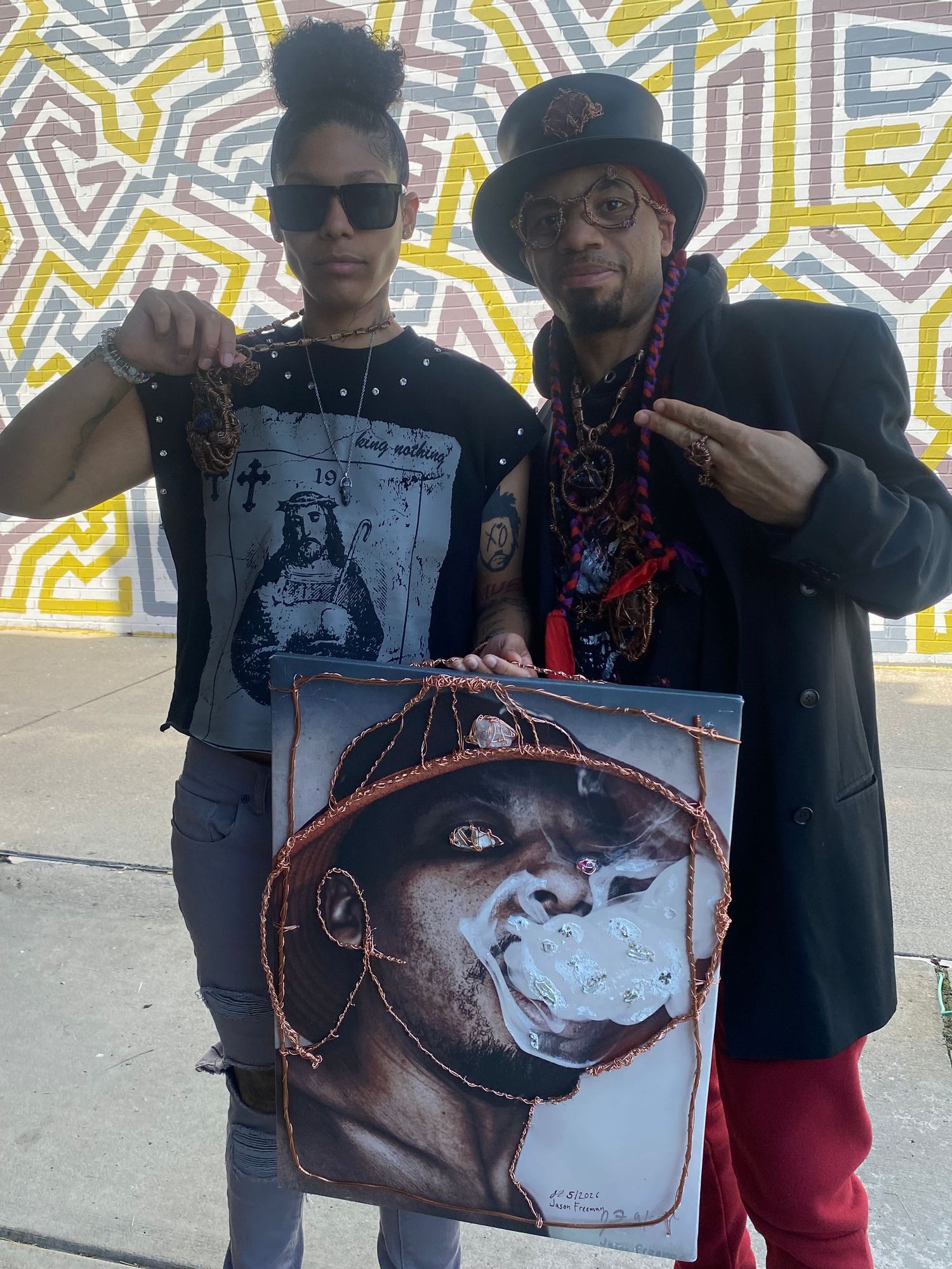 Two artists pose with a vivid portrait painting in front of a colorful geometric mural outdoors.