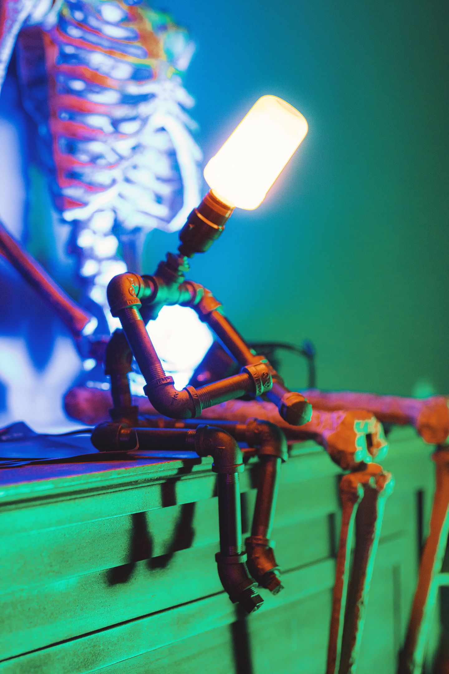 A steampunk pipe figure desk lamp with glowing bulb sits on a shelf with a colorful skeleton display in the background.
