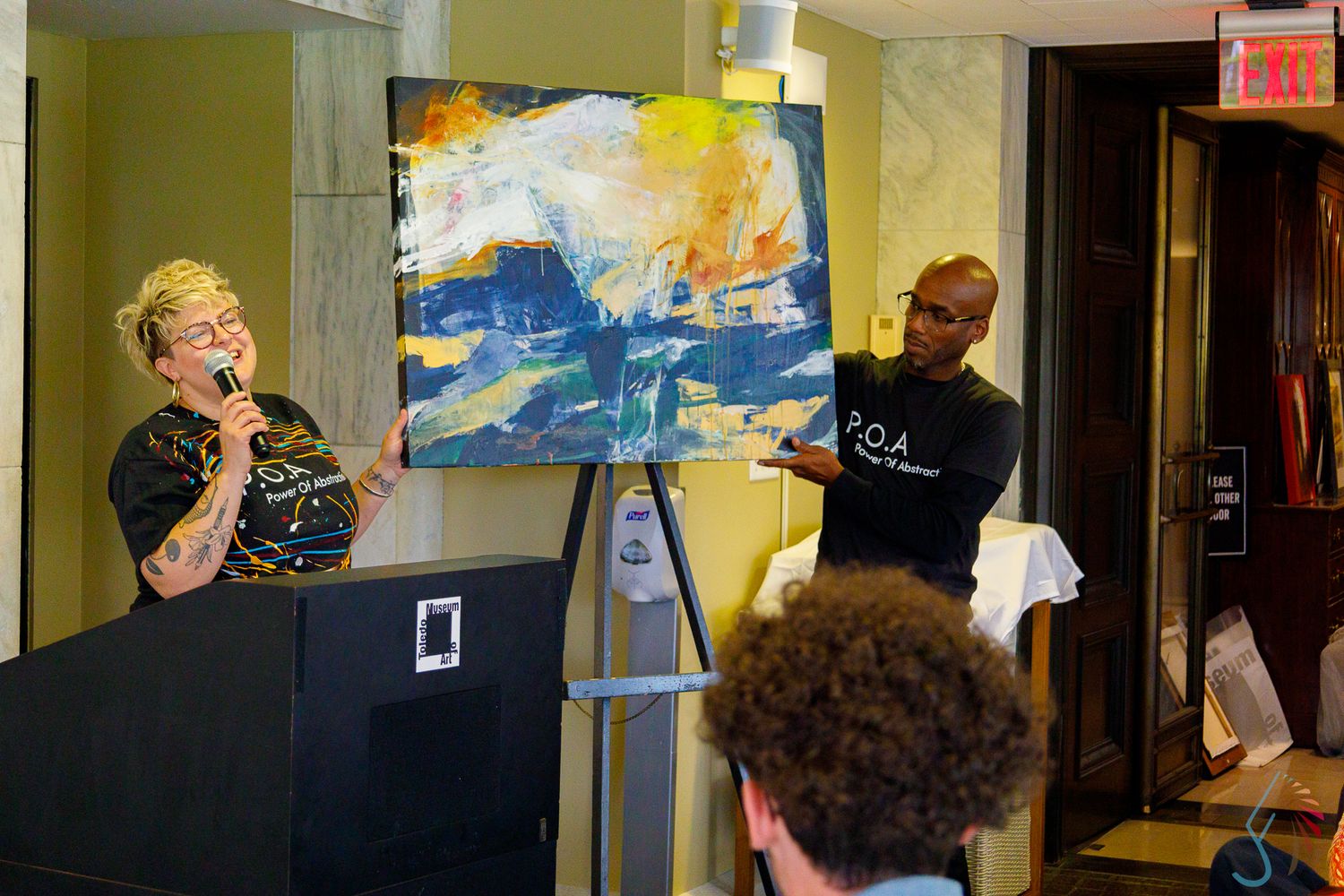 Speaker presents abstract blue and yellow painting on easel at a P.O.A. art event.