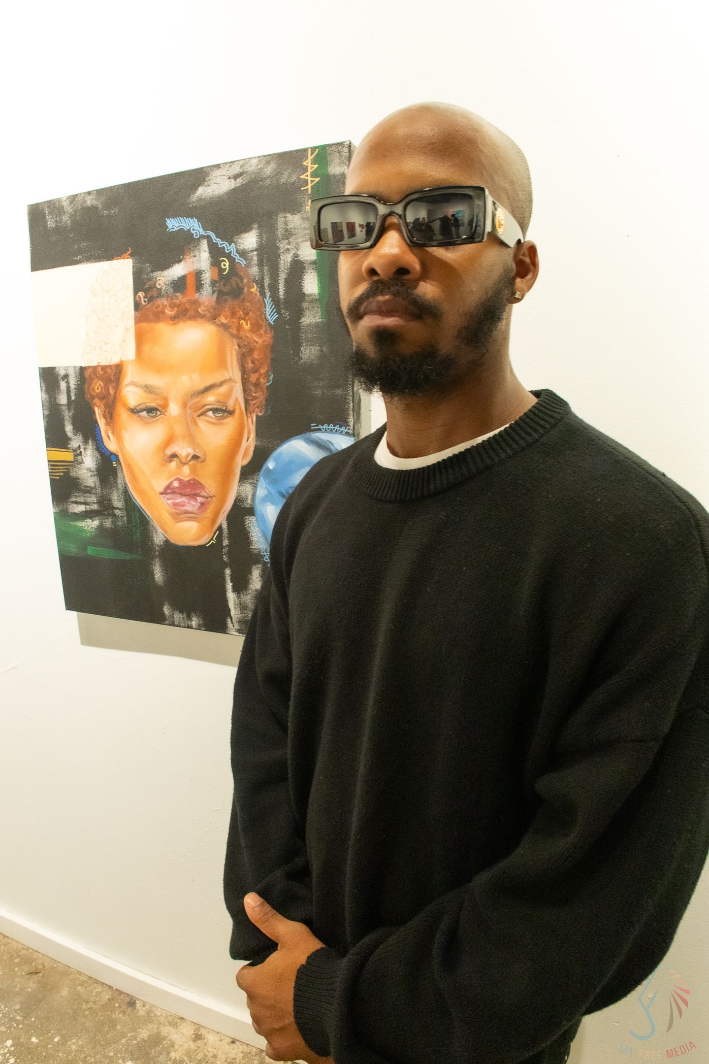 Black man in black sweater and sunglasses poses beside a vibrant portrait painting at an art gallery.