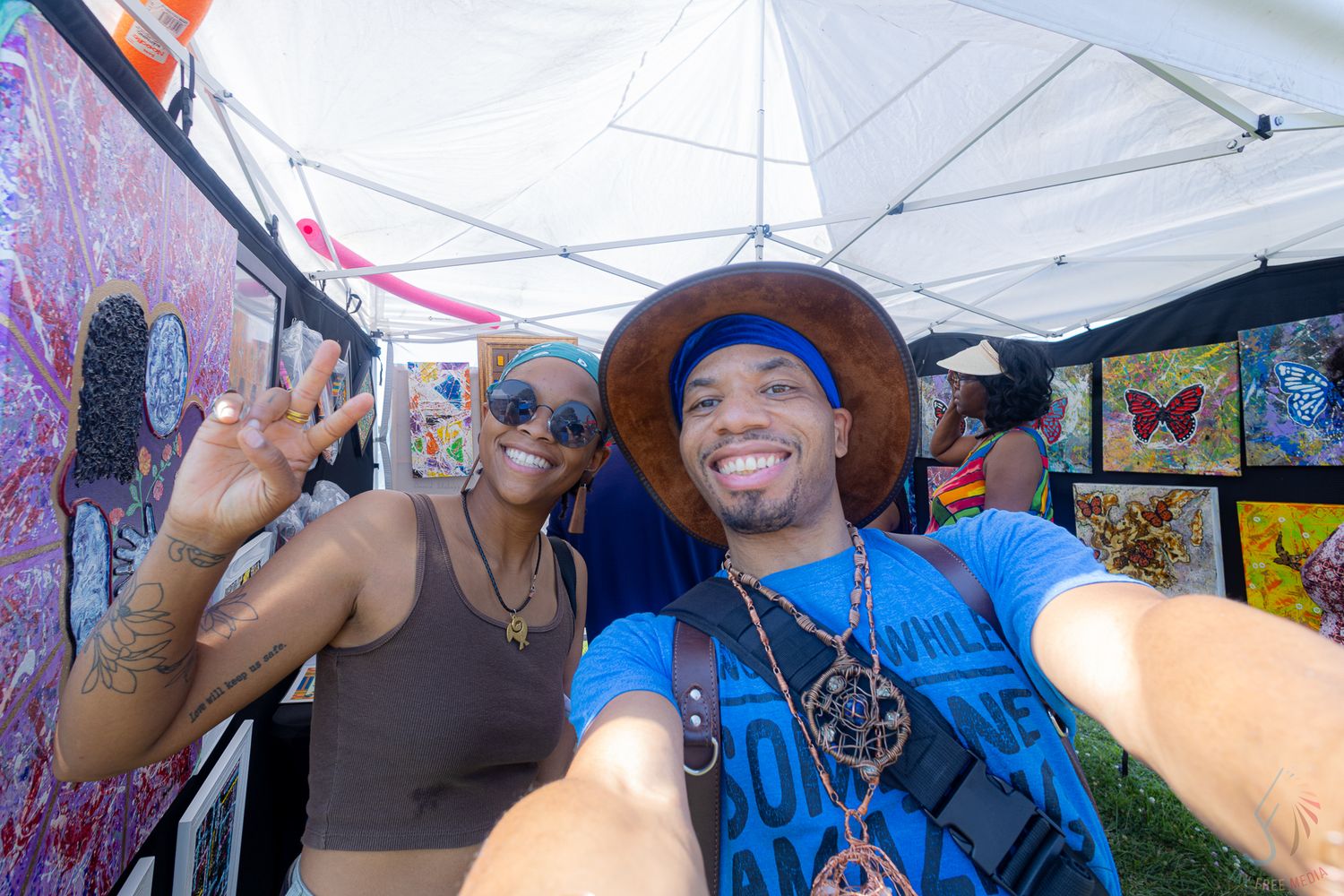 Two friends pose for a selfie at an outdoor art fair, surrounded by colorful paintings under a white tent.