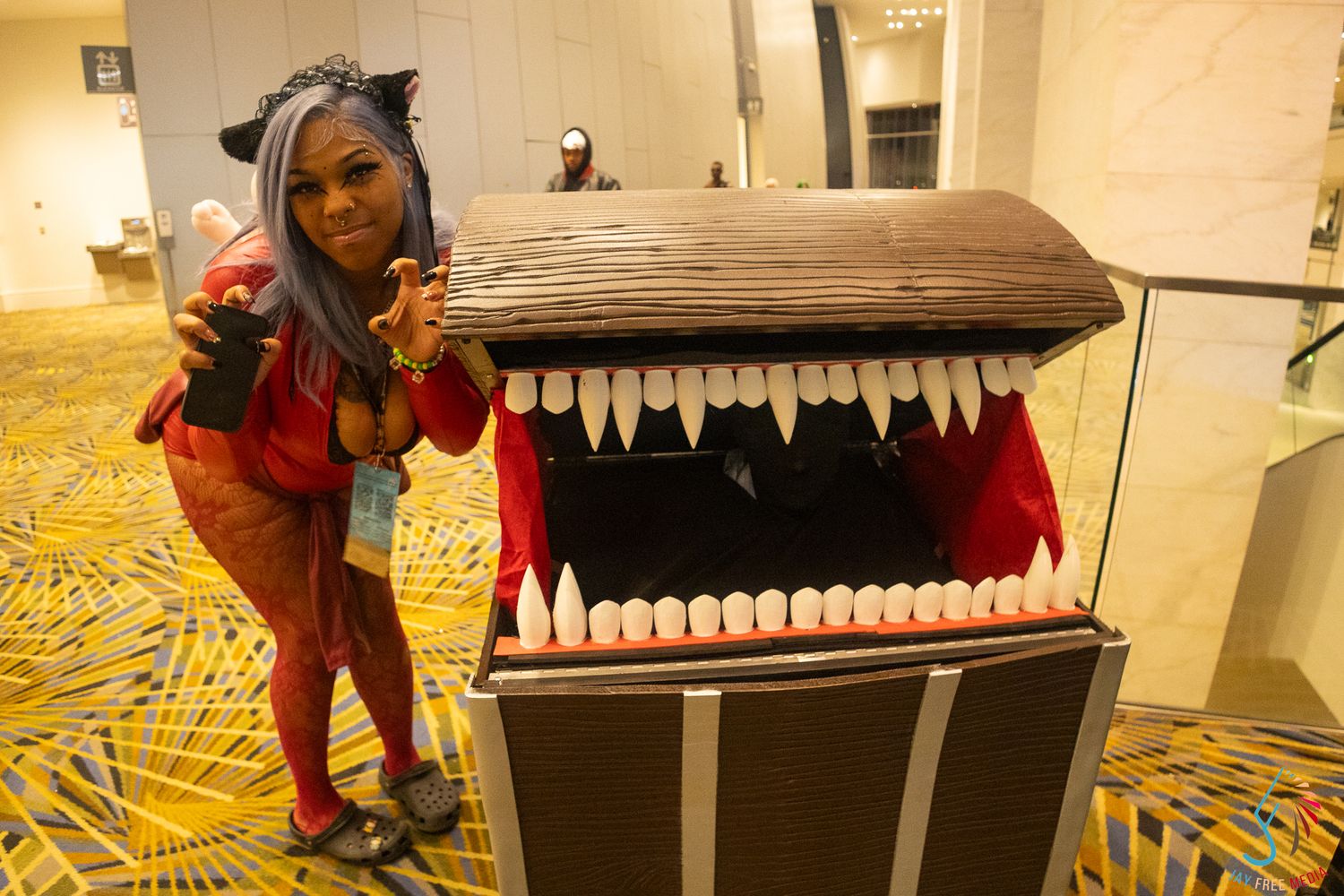 Cosplayer in cat ears poses beside a large mimic chest prop with teeth at a comic convention.