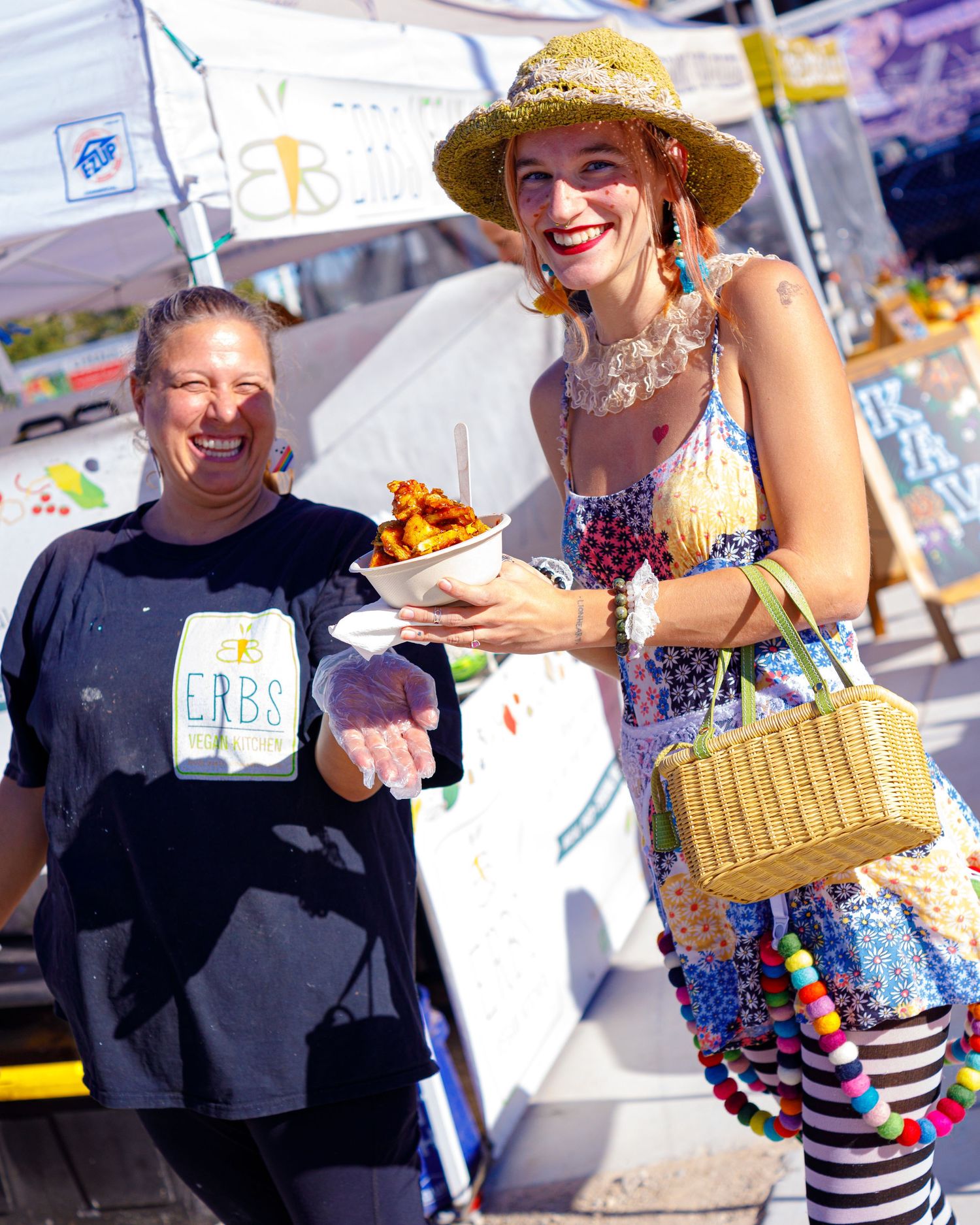Two women smiling and holding food at an outdoor food festival vendor booth.
