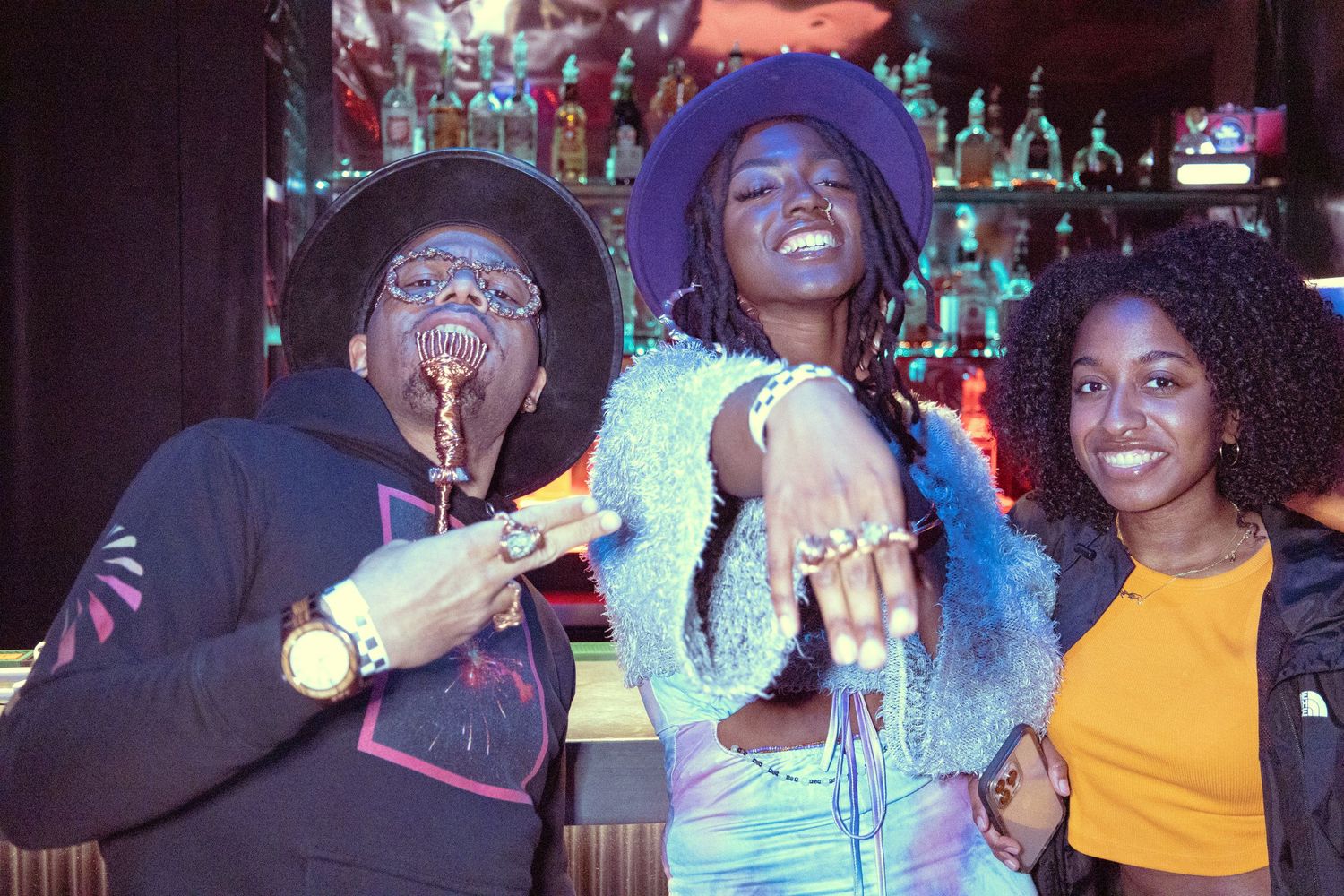 Three friends posing together at a bar, wearing colorful outfits and accessories, smiling at the camera.