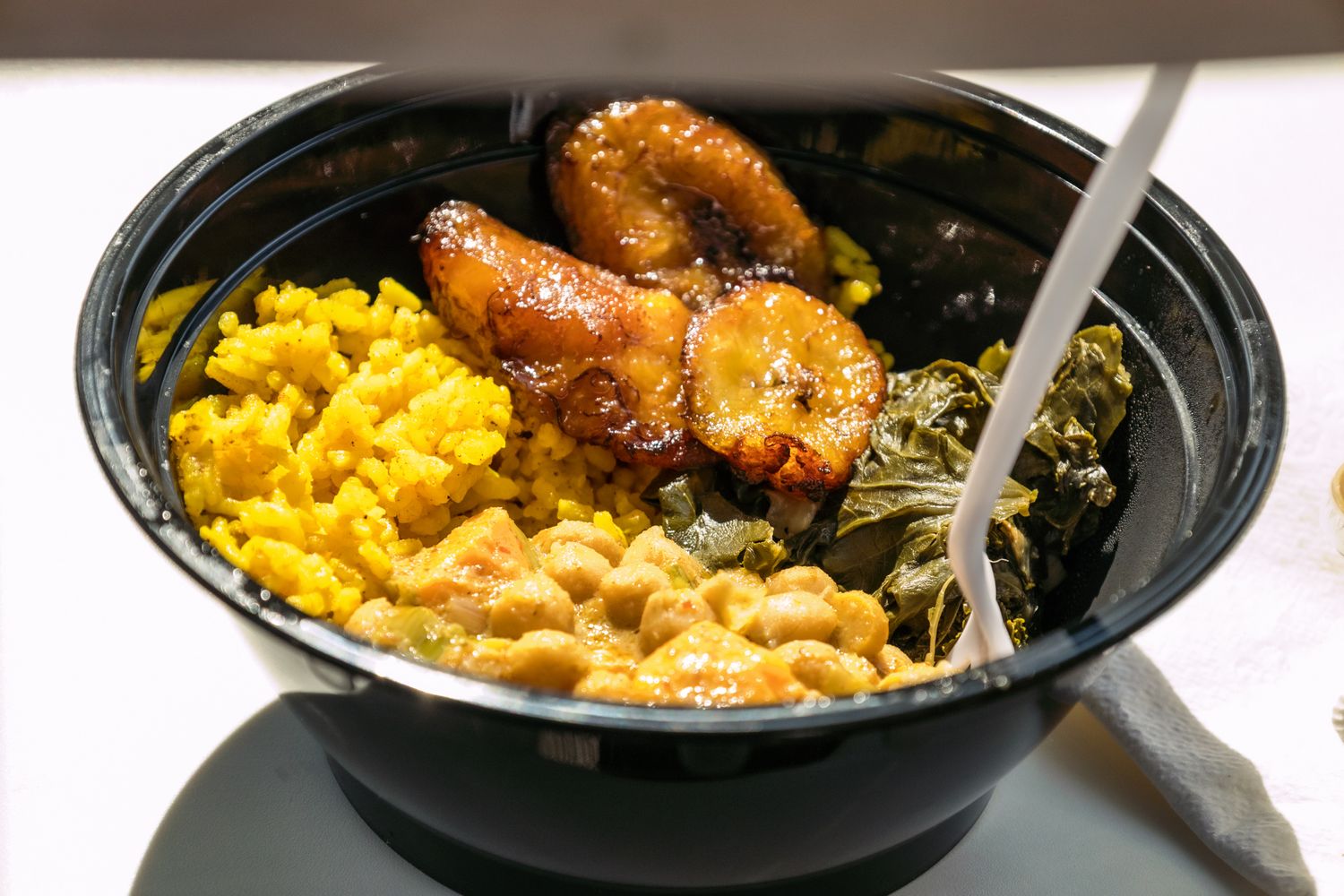Black bowl with yellow rice, collard greens, baked plantains, and beans, a hearty soul food meal.