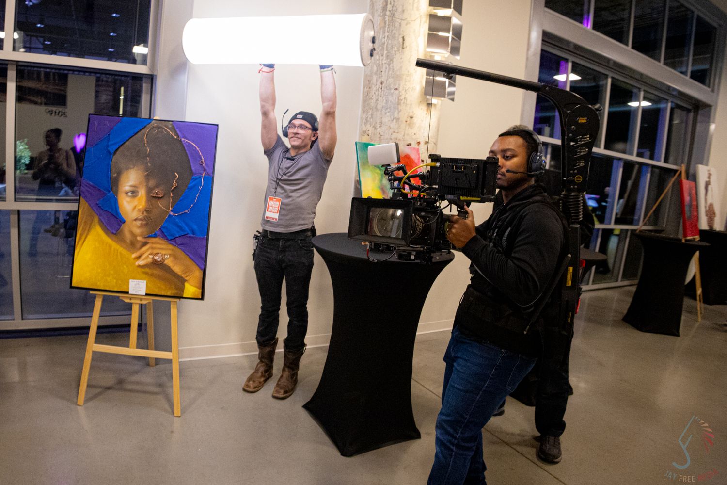 Artist celebrates beside portrait painting at gallery event while videographer films the artwork display.