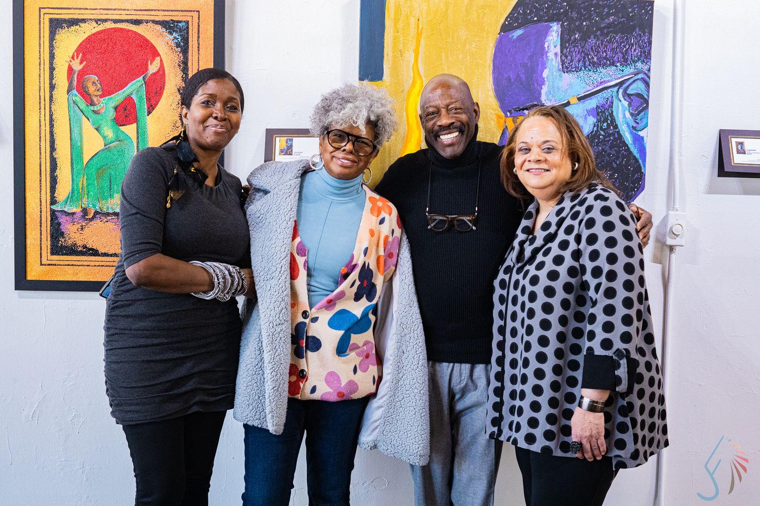 Four people smiling together in an art gallery with colorful paintings displayed on the white walls behind them.