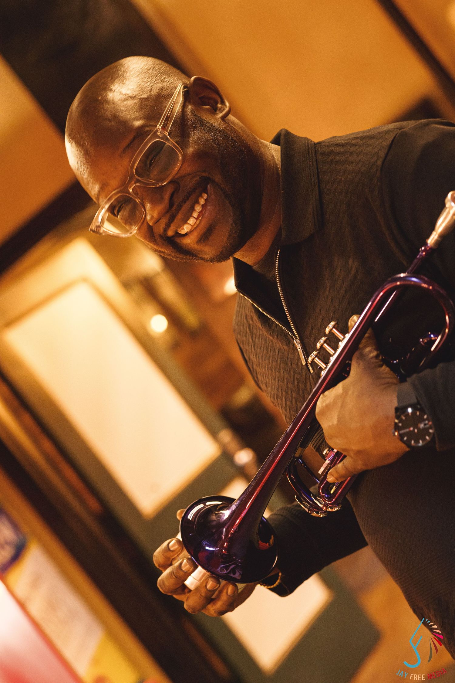 Smiling musician holding a purple trumpet, wearing glasses and a dark jacket, performing in a warmly lit venue.