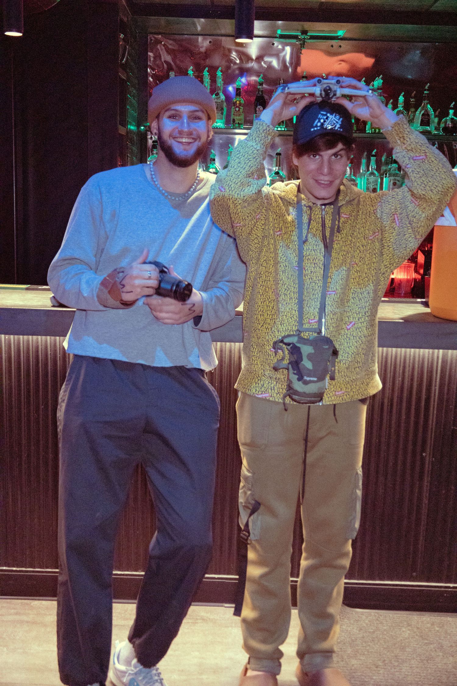 Two young men stand in front of a bar, one holding a camera and the other adjusting a hat on his head.