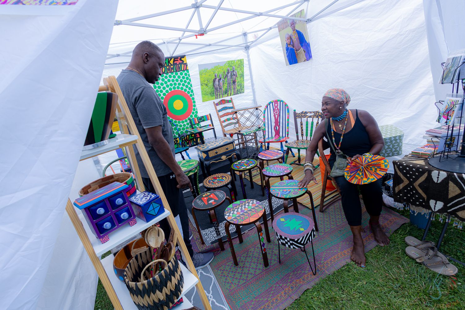 Two people browse colorful African-inspired painted furniture and woven baskets displayed in an outdoor market tent.