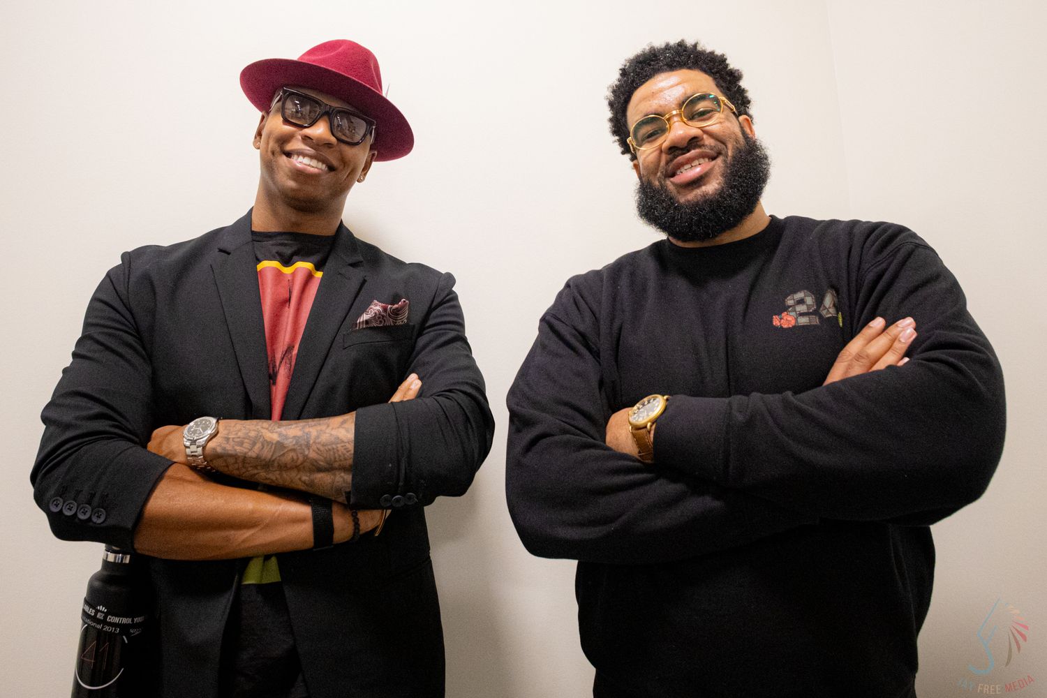 Two smiling men posing with arms crossed, one wearing a red hat and black blazer, the other in a black hoodie.