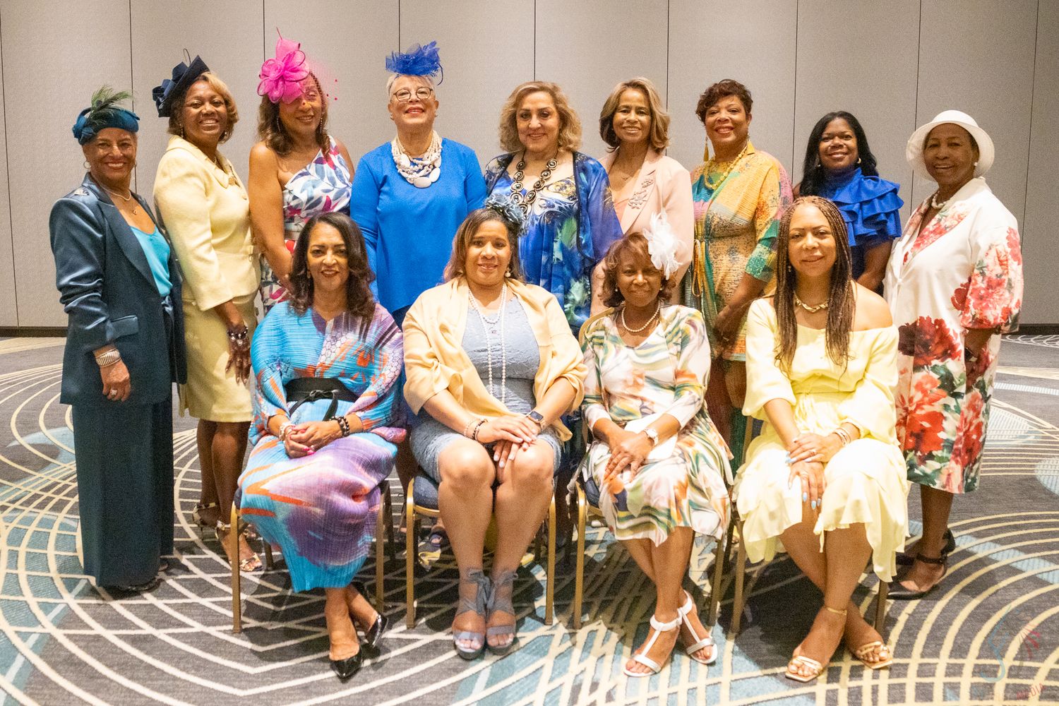 Diverse group of elegantly dressed women in colorful outfits posing together for a formal group portrait indoors.