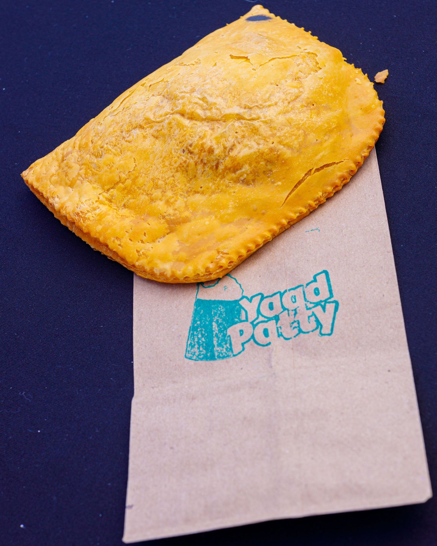 A golden Jamaican beef patty rests on a Yaad Patty branded paper sleeve on a blue surface.