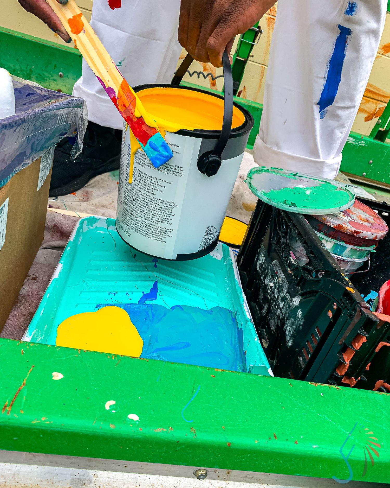 Person mixing bright yellow and blue paint in a white bucket with a brush over a green tray.