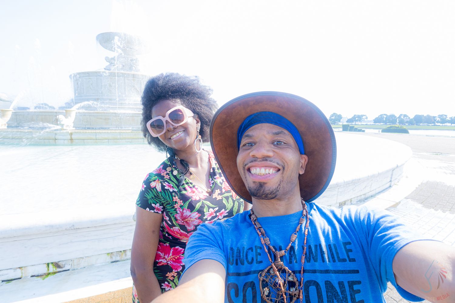 Smiling couple takes a selfie in front of a large fountain on a sunny day.