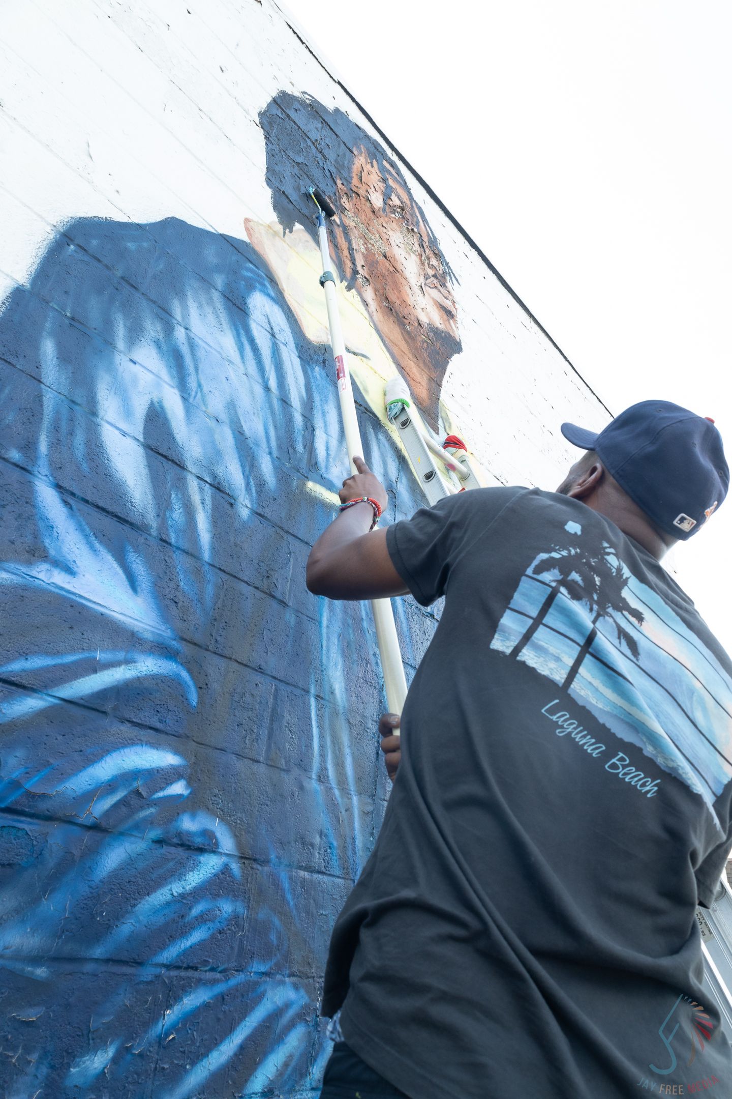 Artist painting a large blue mural on a brick wall outdoors, wearing a Laguna Beach t-shirt and cap.