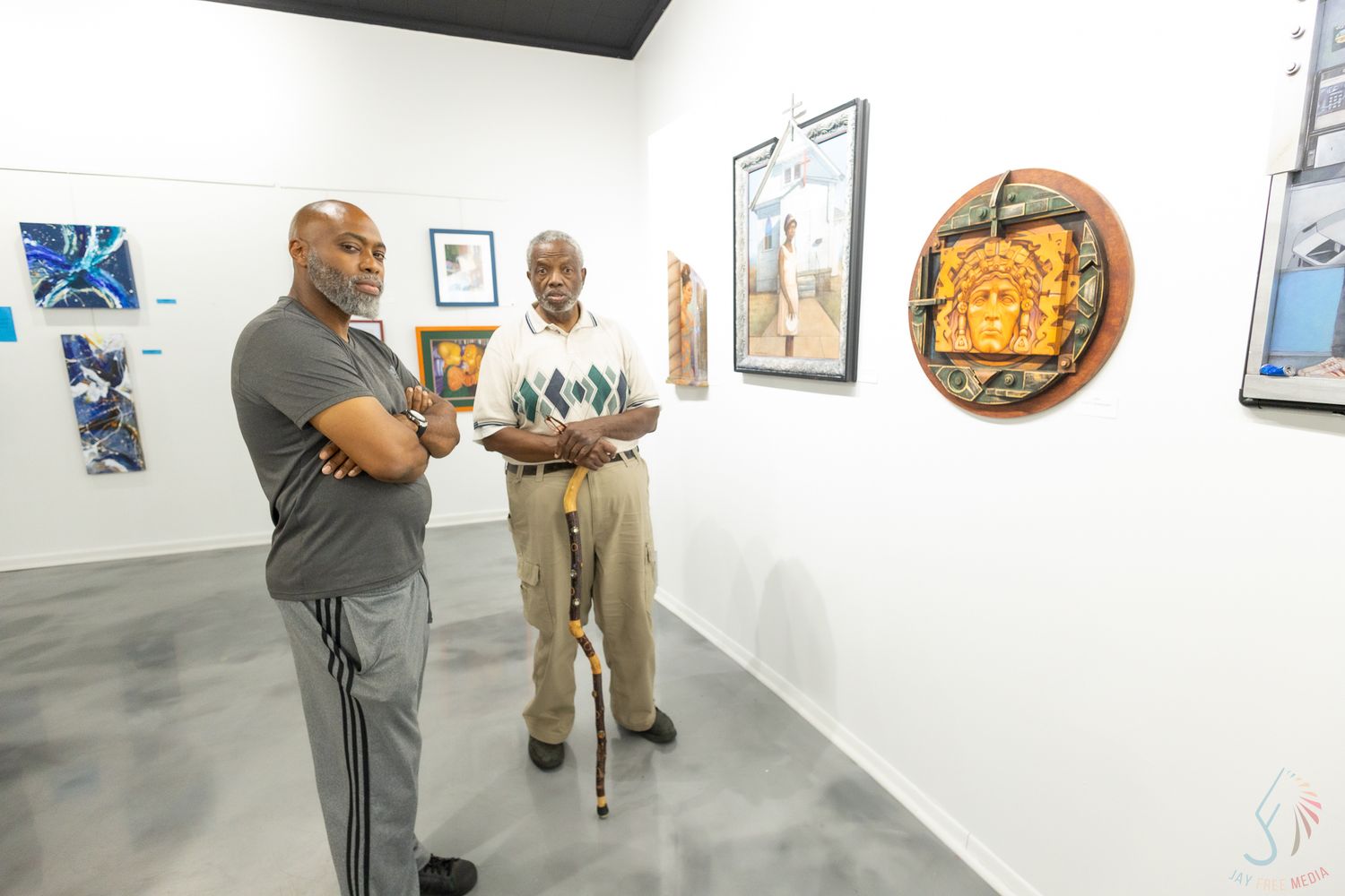 Two men conversing in an art gallery surrounded by colorful framed artwork and circular sculptures on white walls.