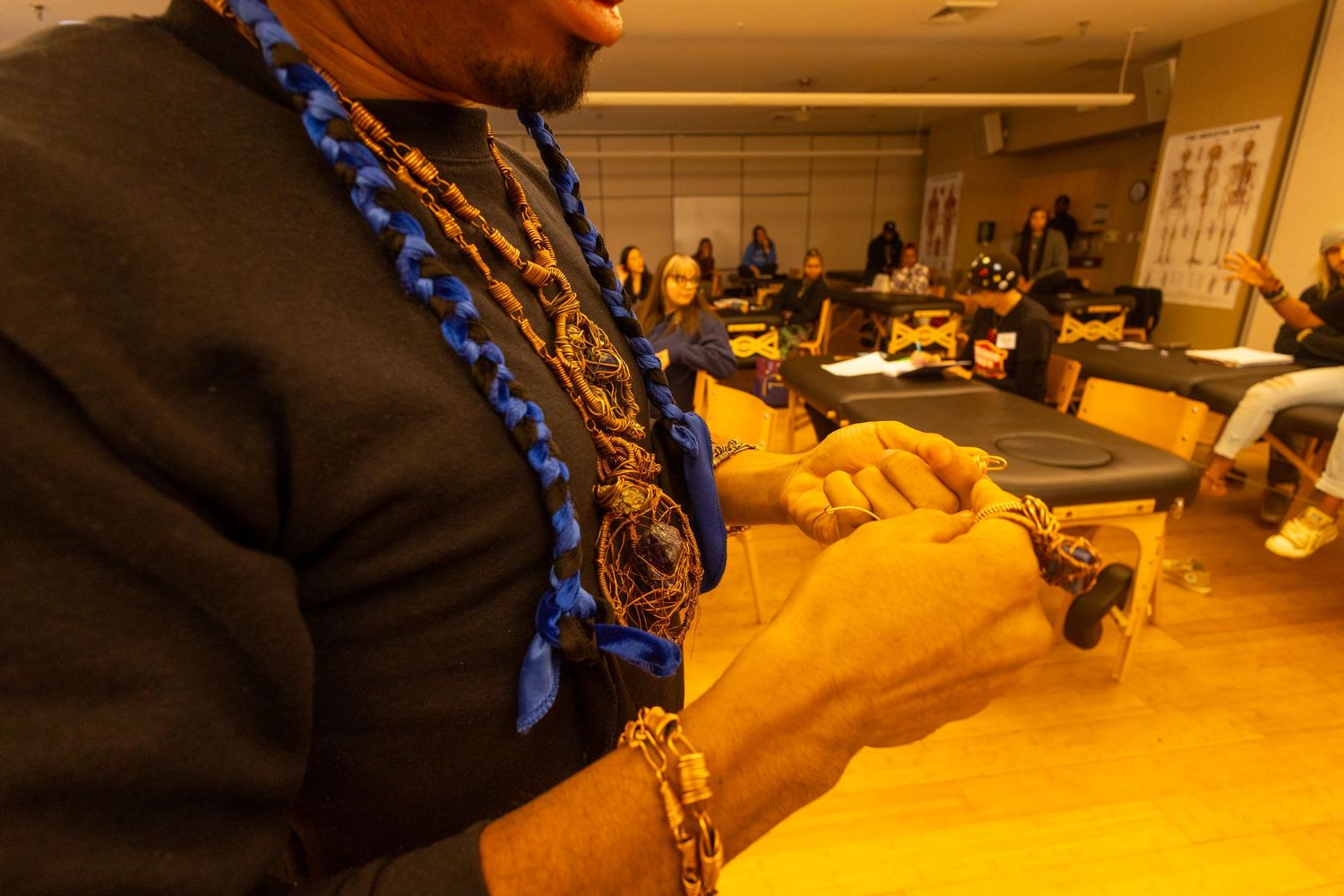 Person wearing colorful beaded necklaces works with hands at a table in a warmly lit community gathering room.