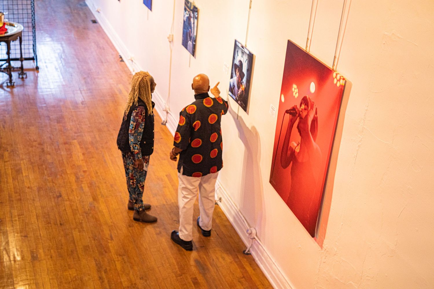 Two visitors view colorful photographs displayed on white walls at an art gallery exhibition.
