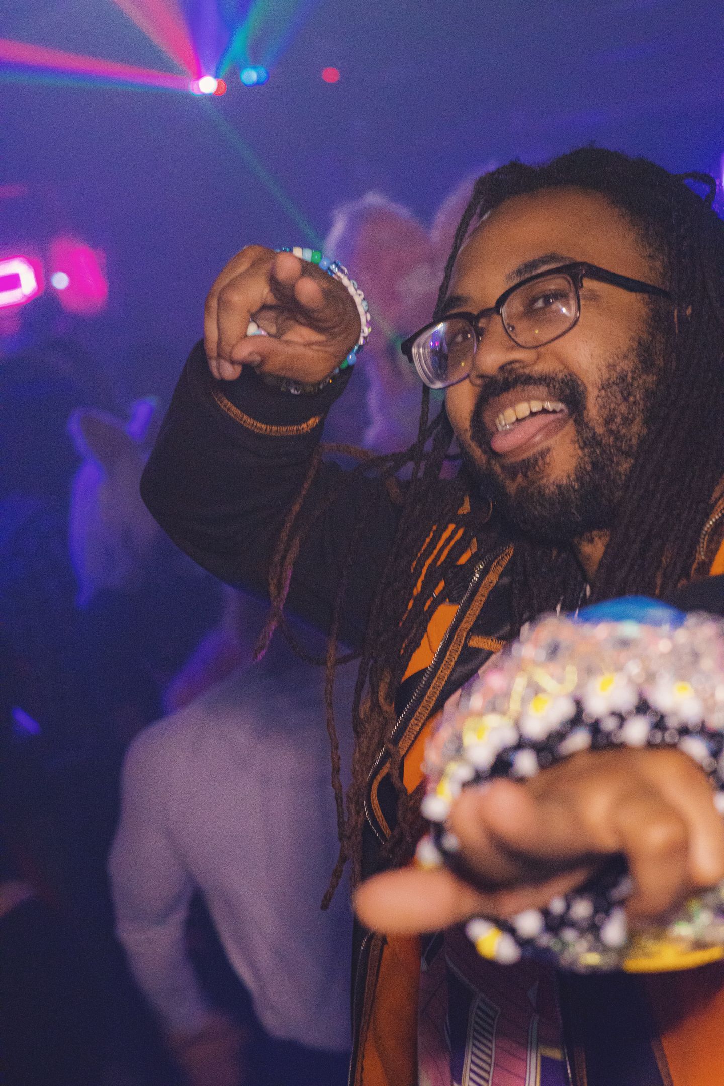 A smiling man with dreadlocks and glasses dancing in a nightclub with colorful laser lights.
