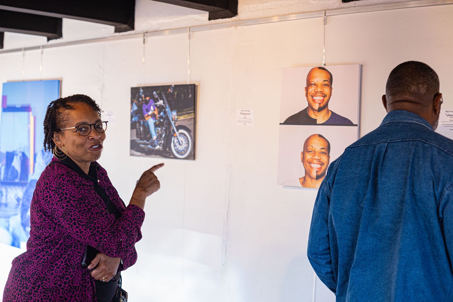 Woman points at portrait photographs displayed on white gallery wall while man looks on.