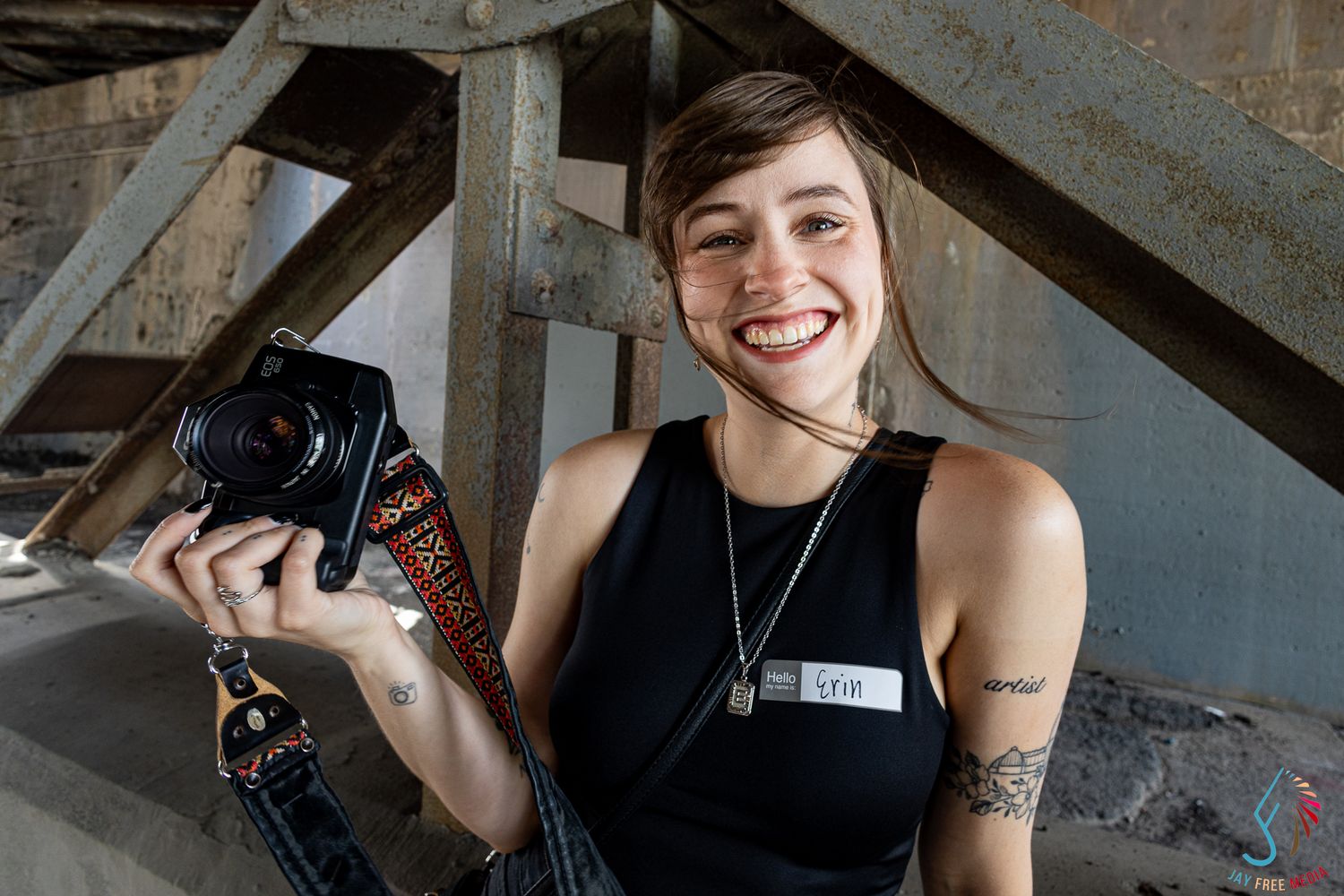 Smiling young woman with tattoos holds a film camera on a colorful strap at an indoor event.