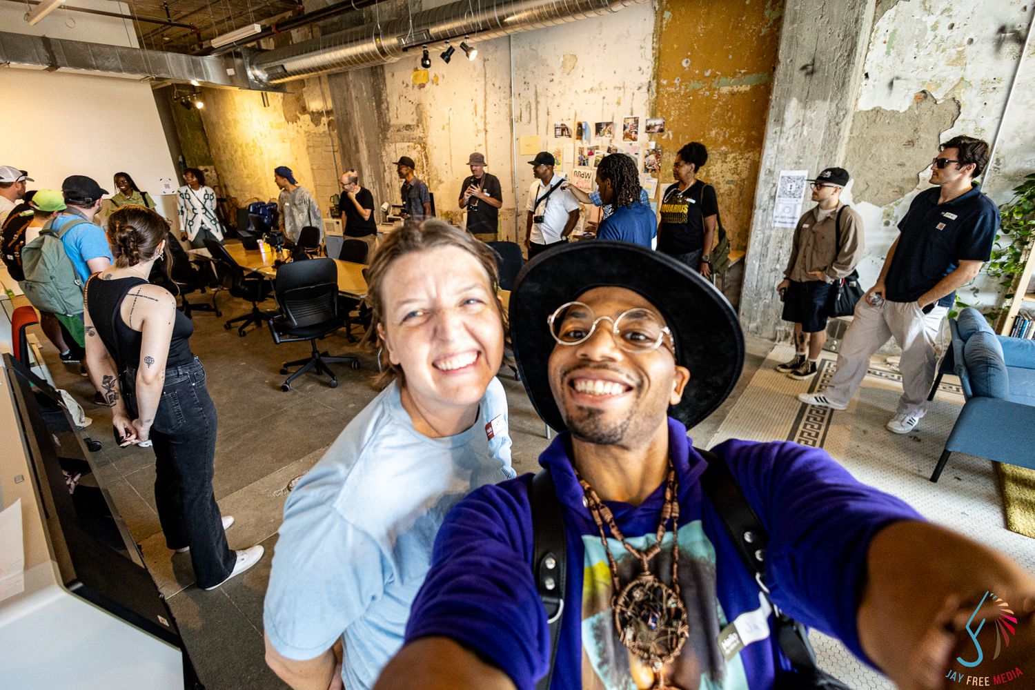 Two people take a smiling selfie at a lively indoor creative event with guests mingling in the background.