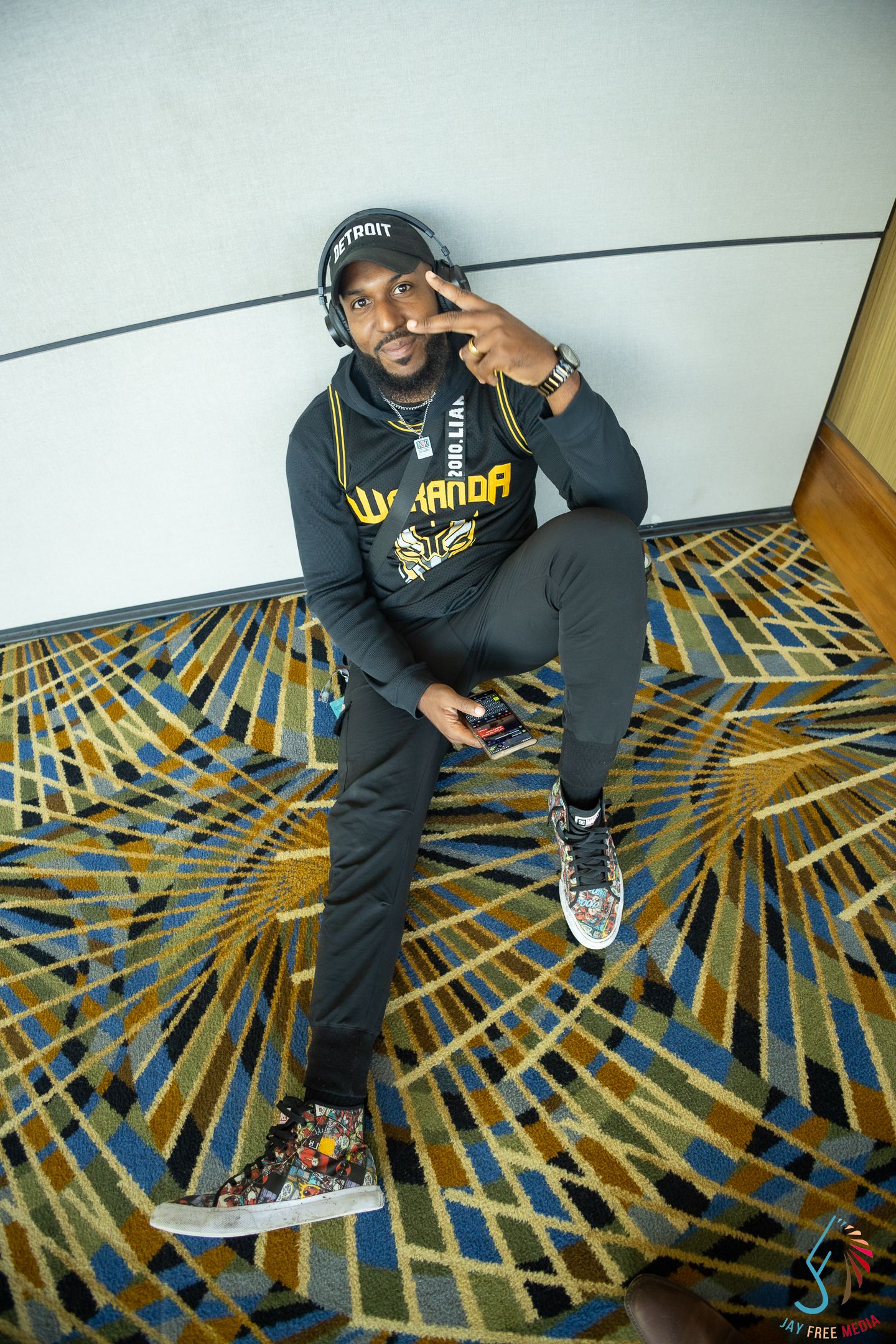 Man in Wakanda hoodie and Detroit cap sits on patterned carpet, flashing peace sign while wearing headphones.