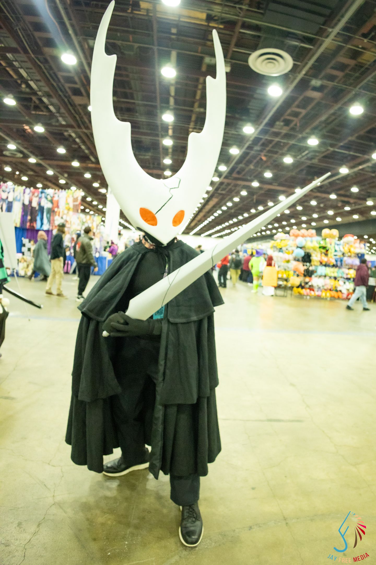 Cosplayer dressed as Hollow Knight's Hornet character with white mask and black cape at comic convention.