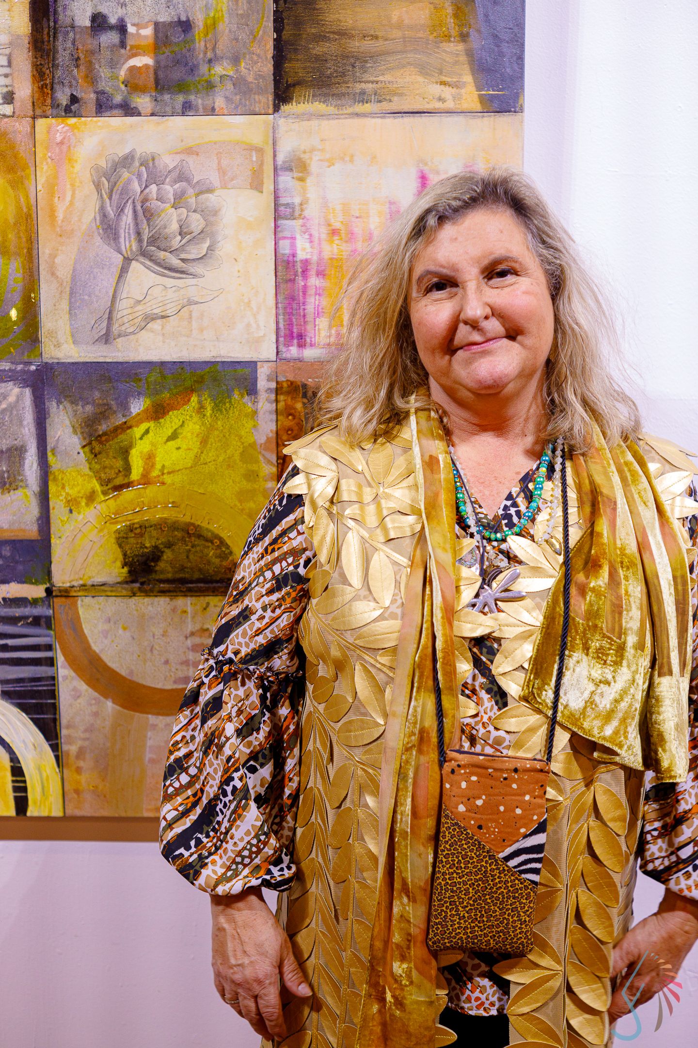 Smiling woman in gold outfit stands before colorful abstract paintings featuring floral motifs in yellow and gray.