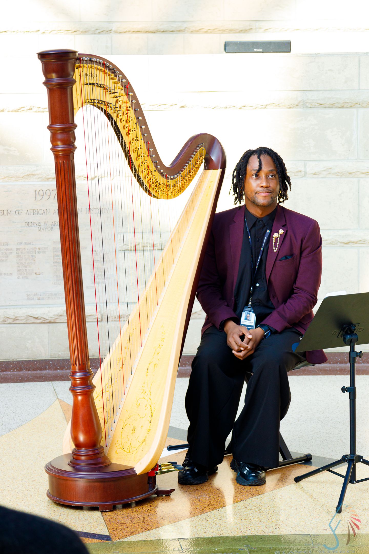Young musician in burgundy blazer poses beside a large concert harp in an elegant marble hall.