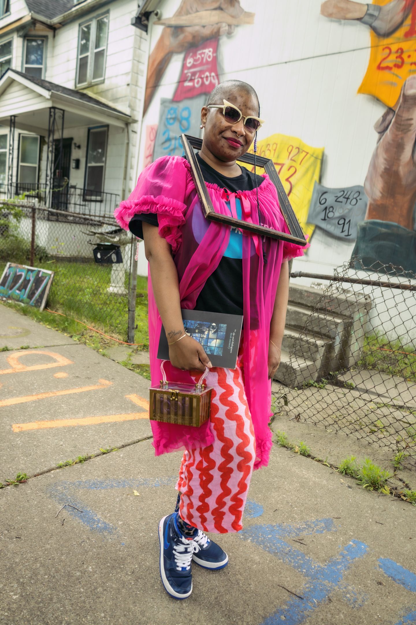 Woman in vibrant pink dress with sunglasses holds artwork outdoors near colorful mural in urban neighborhood.