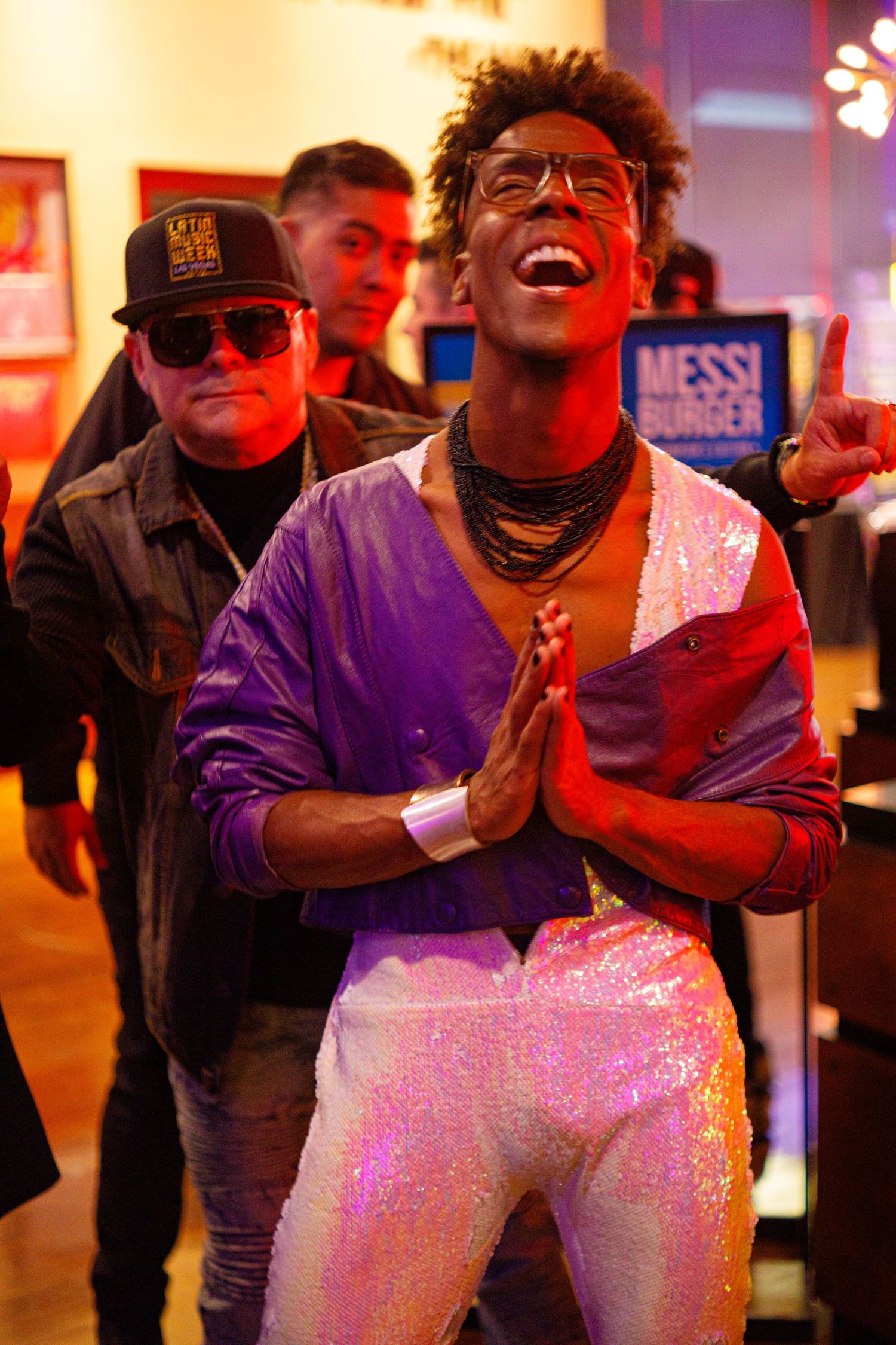 A joyful man in sequined white pants and purple jacket poses with prayer hands at a vibrant indoor event.