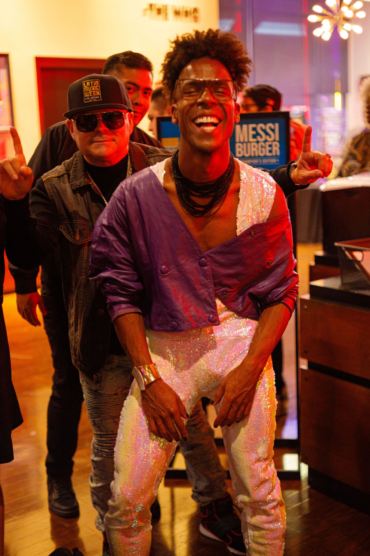 Smiling man in sequin pants holds a Messi Burger sign at a lively event.