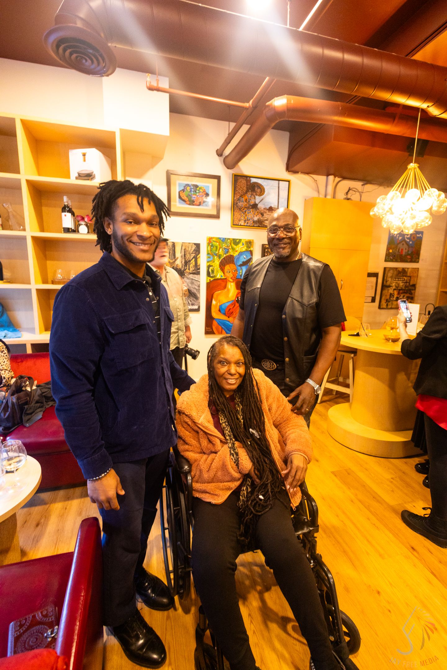Three people smiling in a cozy art-filled lounge, one seated in a wheelchair, surrounded by vibrant artwork.