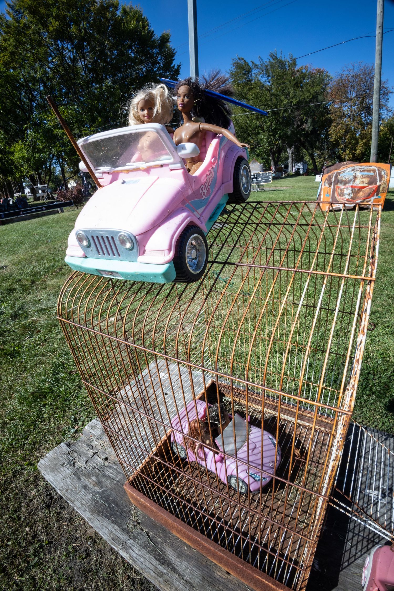 Two Barbie dolls in a pink toy Jeep balanced on top of a rusty vintage birdcage outdoors in a park.