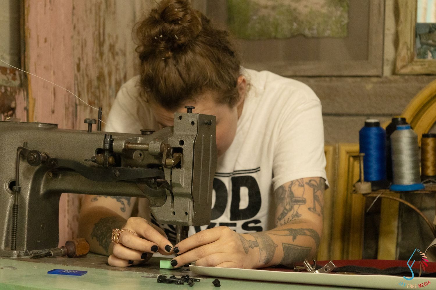Tattooed woman in white t-shirt focused on sewing with industrial machine in a rustic workshop setting.